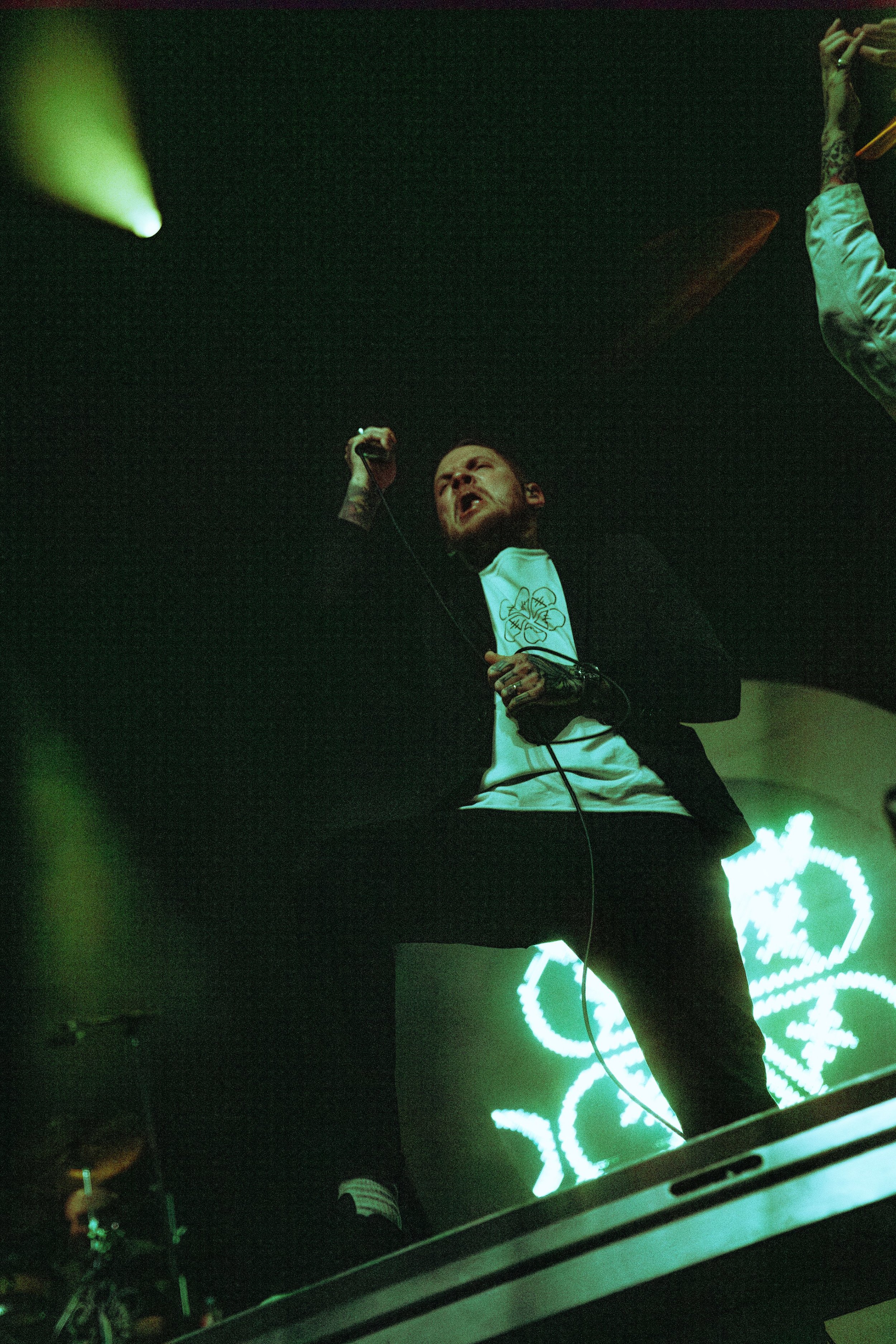 A man singing into a microphone on a dark stage with green lighting and neon heart and flower shapes in the background.