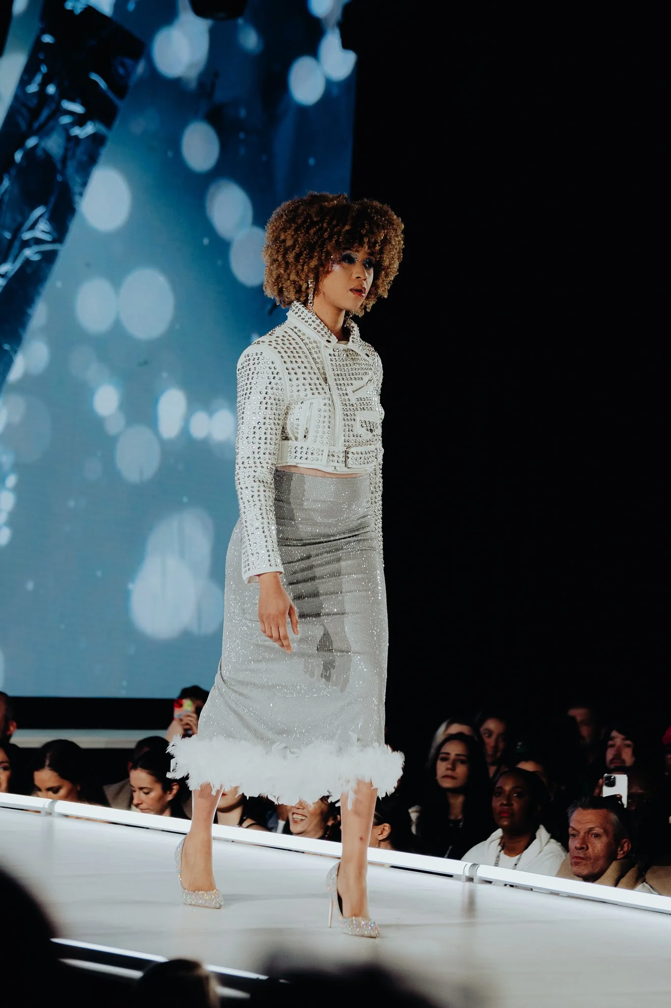 A woman in a white, rhinestone-studded jacket and skirt with feathered hem walks on stage during a fashion show, with a background of blue bokeh lights and a crowd watching.