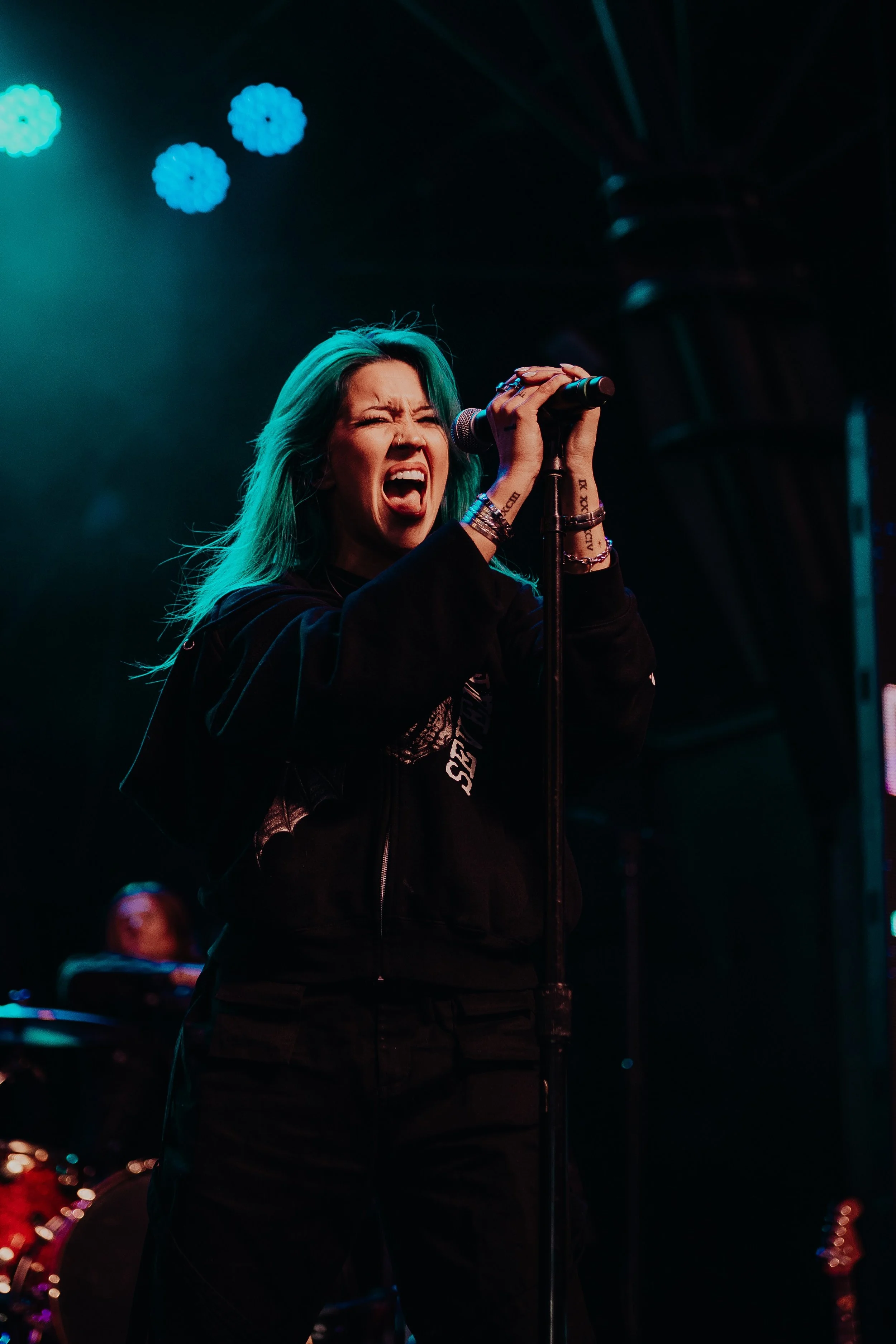 A woman with teal hair passionately singing into a microphone on a stage, with colorful lights and a keyboard player in the background.