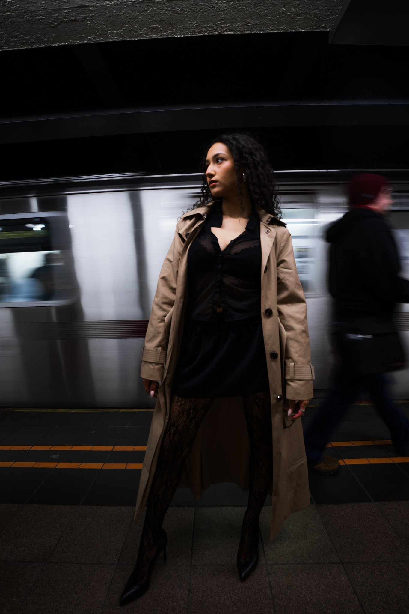 A woman with dark curly hair wearing a beige trench coat, a black lace dress, and black high heels, standing on a subway platform with a moving train behind her. A man in black coat and red hat is blurred in the background.