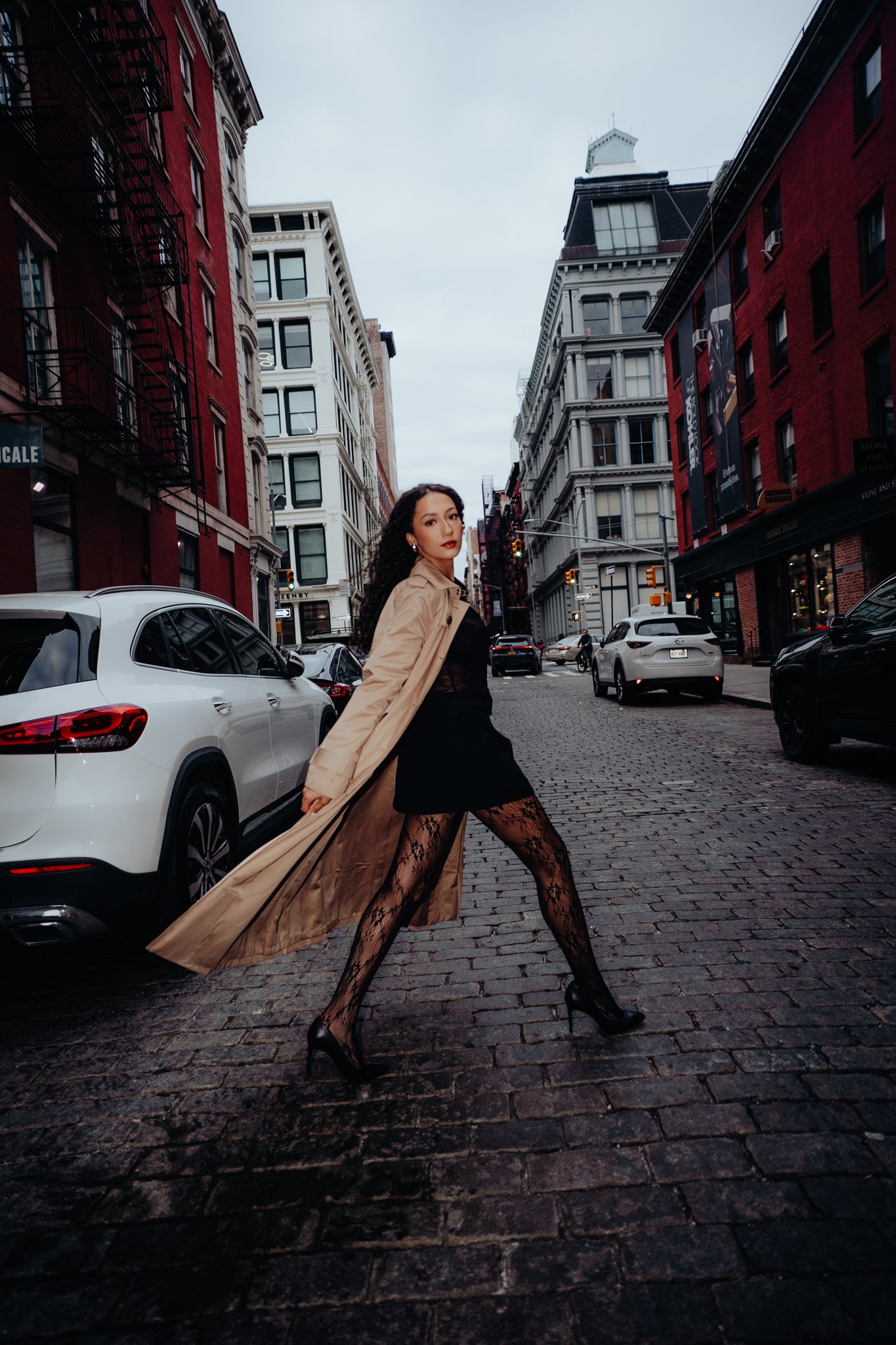 A woman with curly black hair and red lipstick posing on a cobblestone street in an urban area with red and white buildings. She is wearing a black dress, lace stockings, high heels, and a tan trench coat, striking a confident pose.