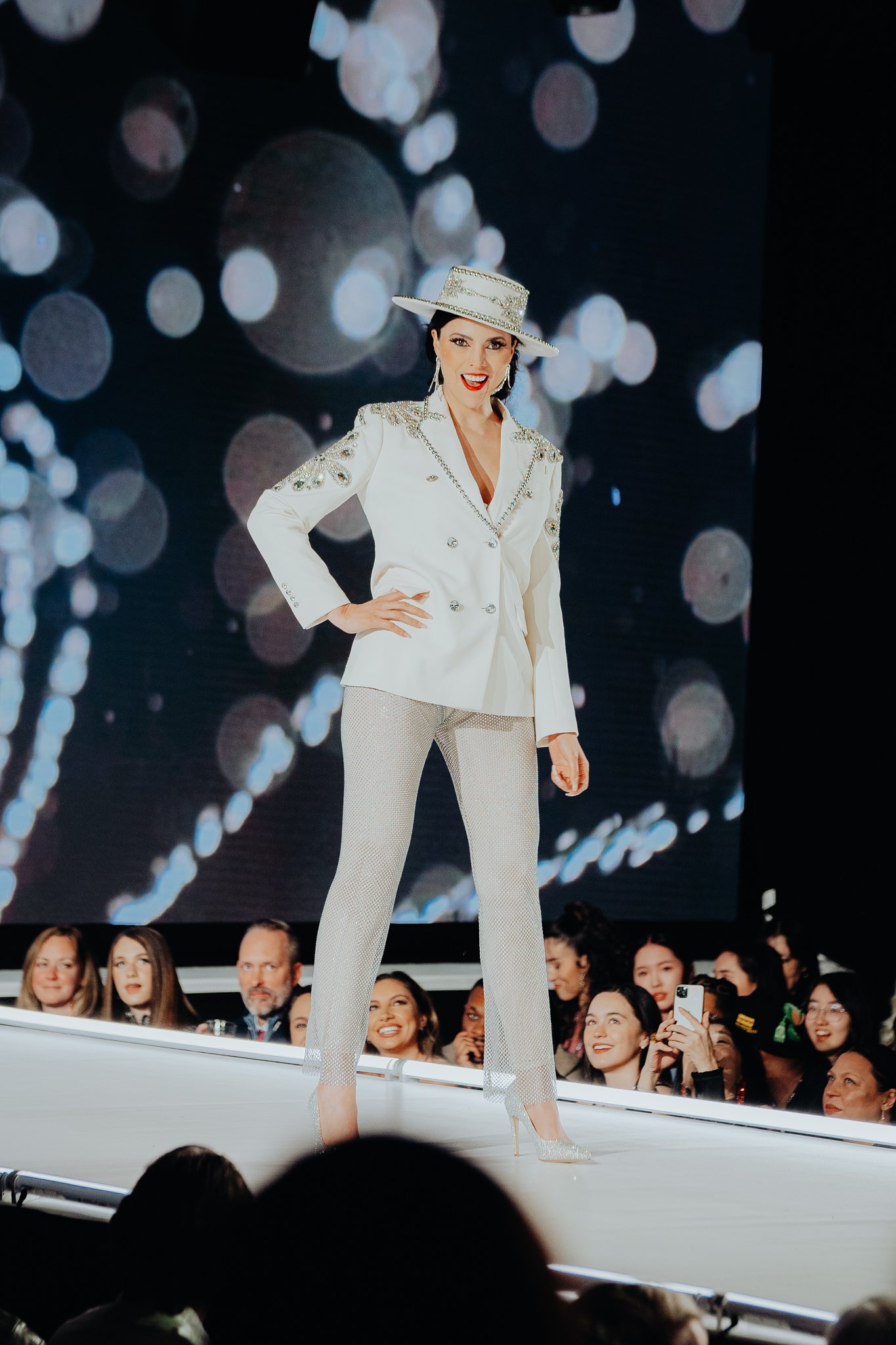 A woman in a white suit with embellishments, a wide-brimmed hat, and high heels walking on a runway with an audience watching and taking photos.