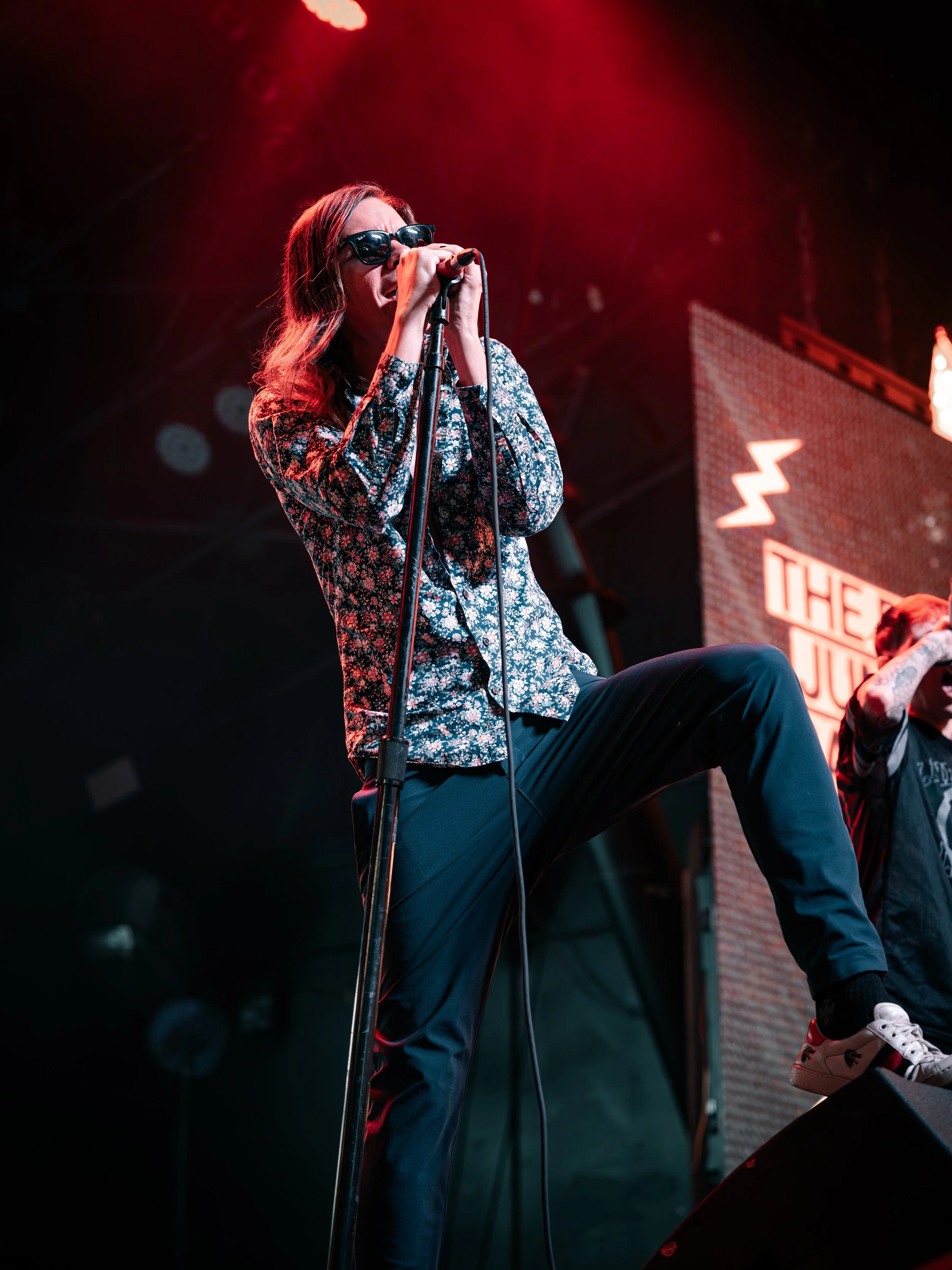 A woman with long hair wearing sunglasses, a floral shirt, and black pants is singing into a microphone during a live performance on stage with red lighting.