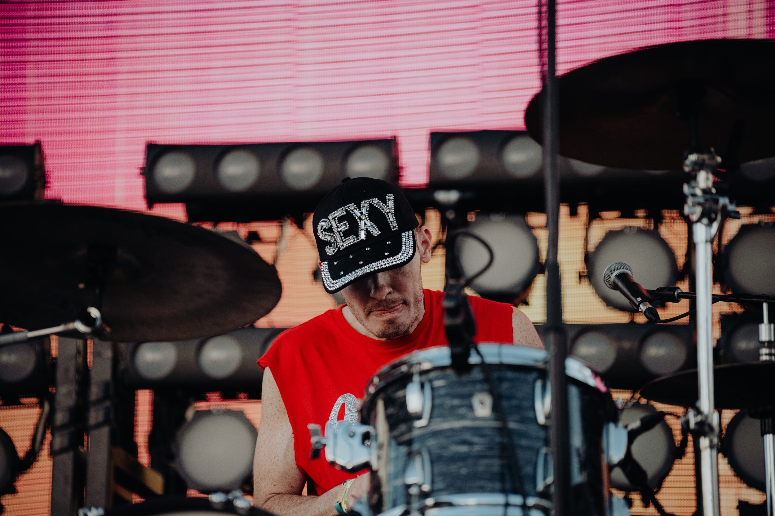 A musician wearing a black cap with the word 'SEXY' encrusted with rhinestones, playing drums on stage during a live performance with a large pink LED screen in the background.