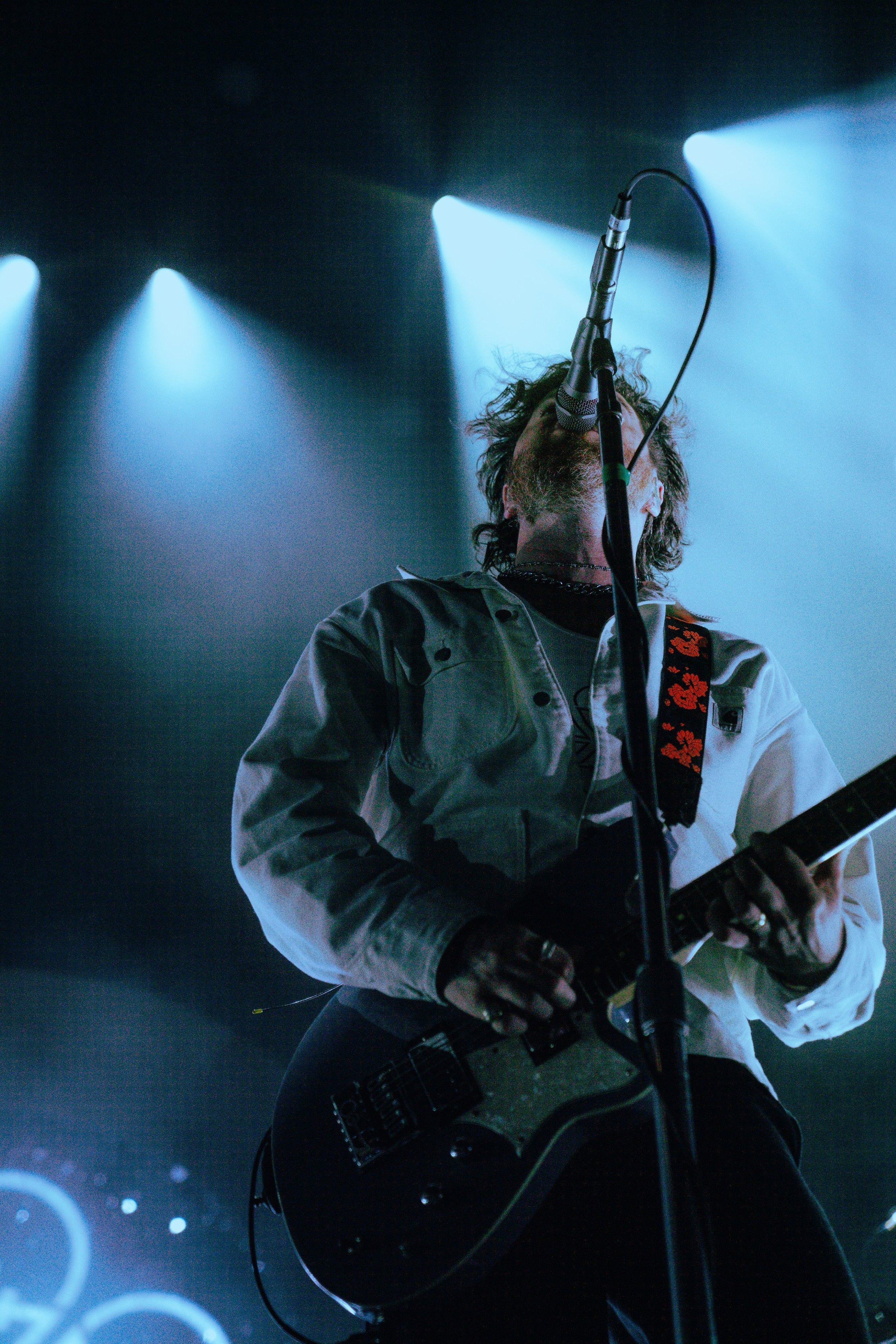 A musician with curly hair playing an electric guitar on stage. The person is singing into a microphone with stage blue lights illuminating the background.