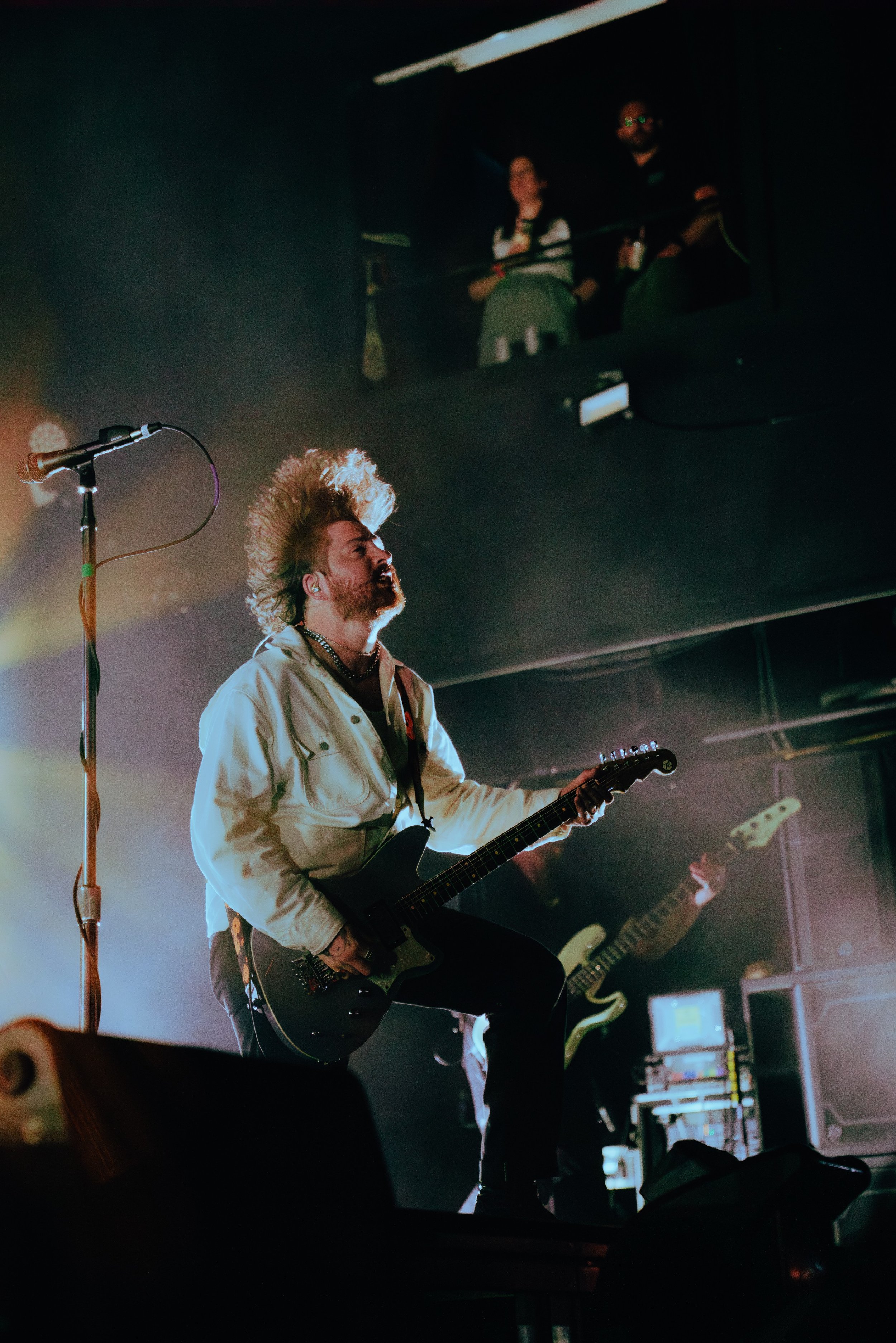 A musician with long, curly hair and a beard playing an electric guitar on stage, with a microphone stand nearby and a band member in the background. The scene is dimly lit with stage lights.