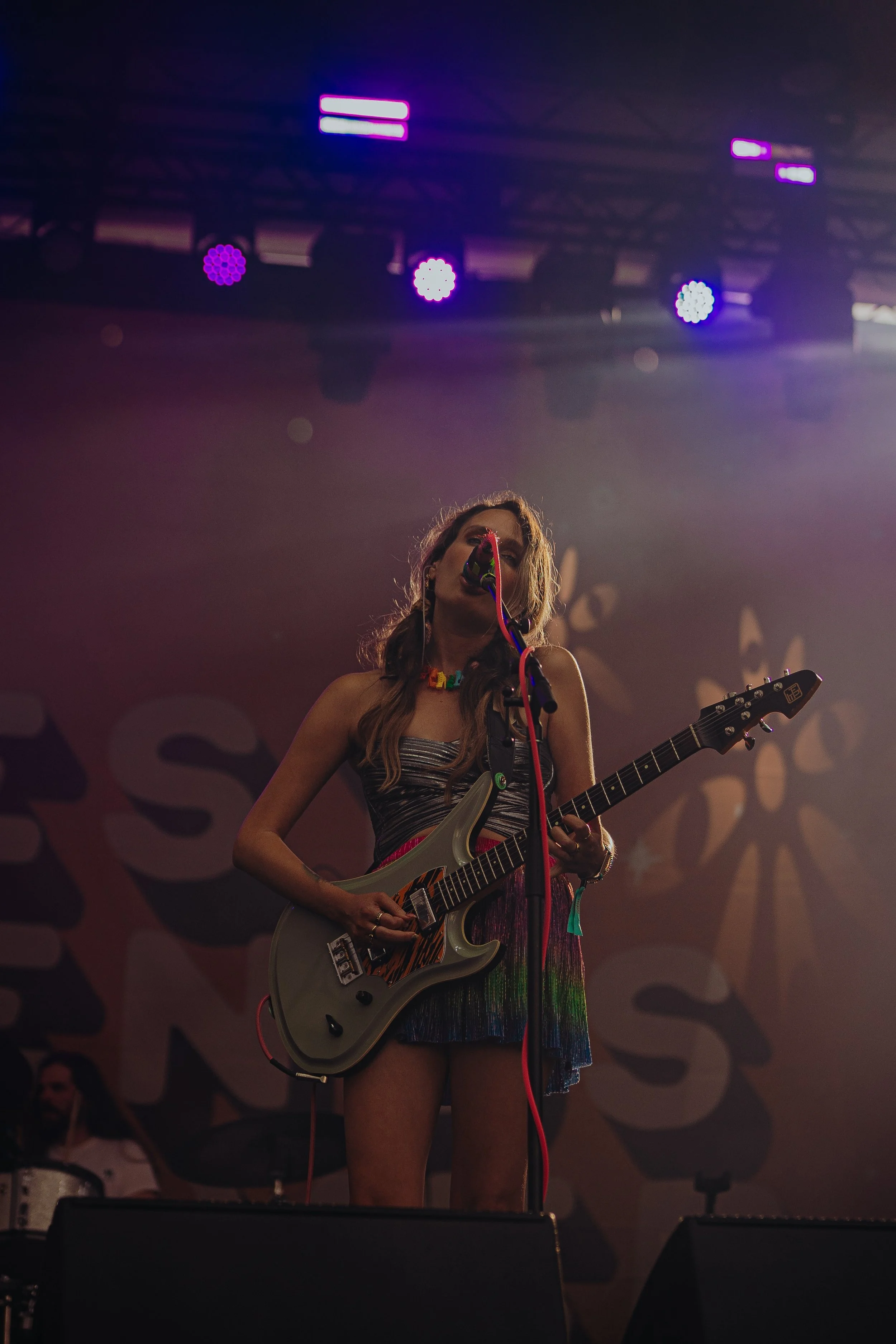 Female musician with long hair singing into a microphone, playing an electric guitar on stage with colorful lights overhead.