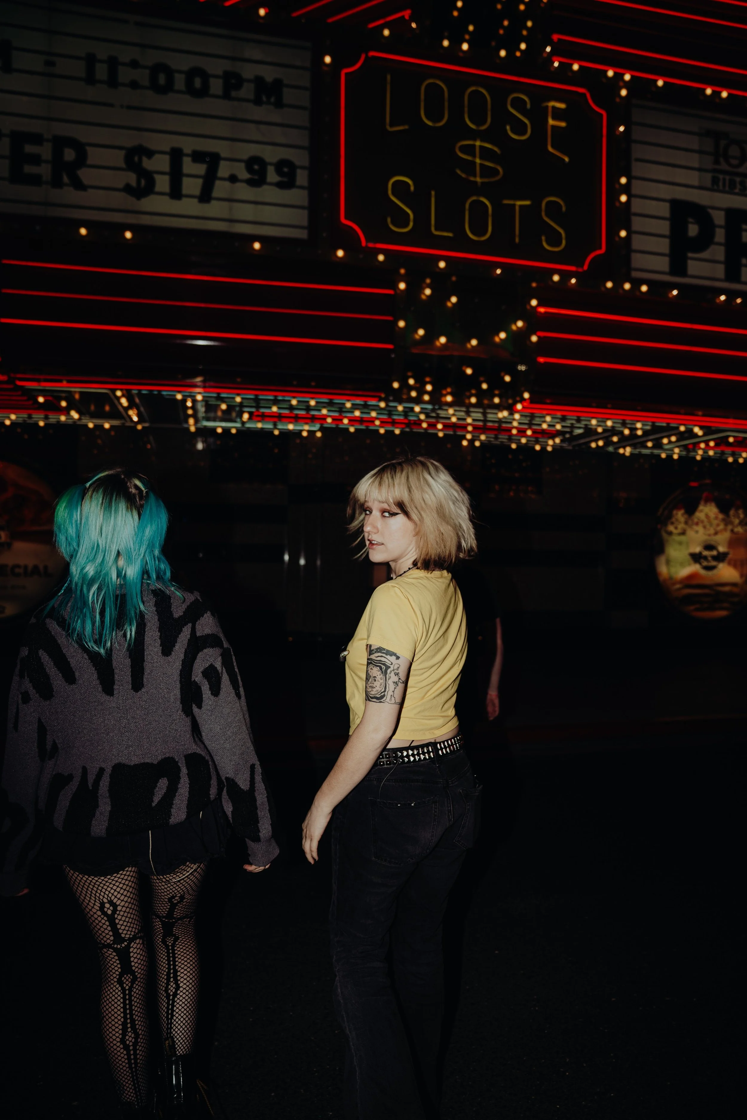 Two young women standing outside a theater at night, illuminated by neon lights and surrounded by marquee signs. One woman has teal hair and fishnet stockings, while the other has blonde hair, tattoos on her arm, and wears a yellow shirt and black je