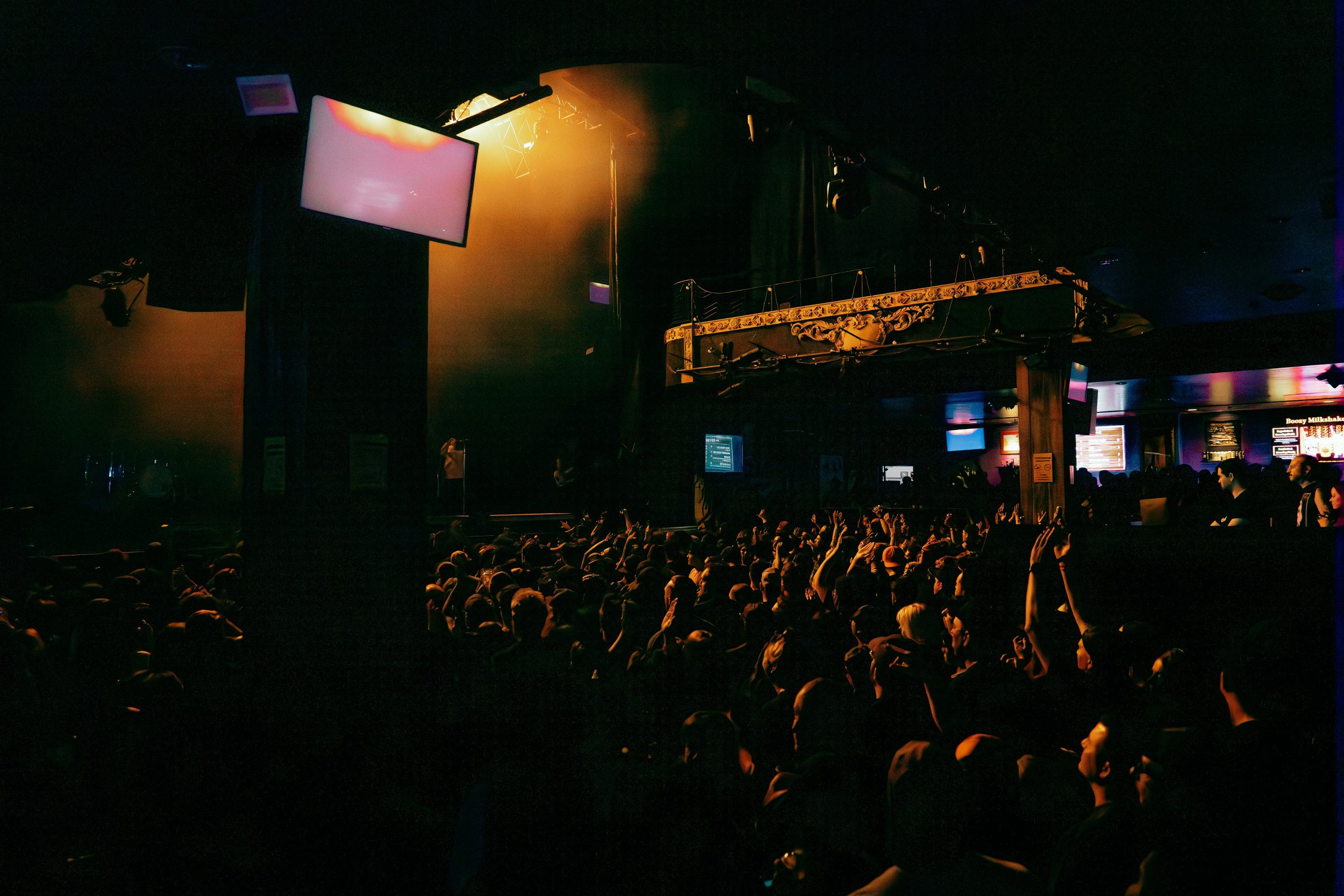 A crowd of people sitting in a dark theater or concert venue, looking towards a stage with screens and lighting, with some of the audience raising their hands.