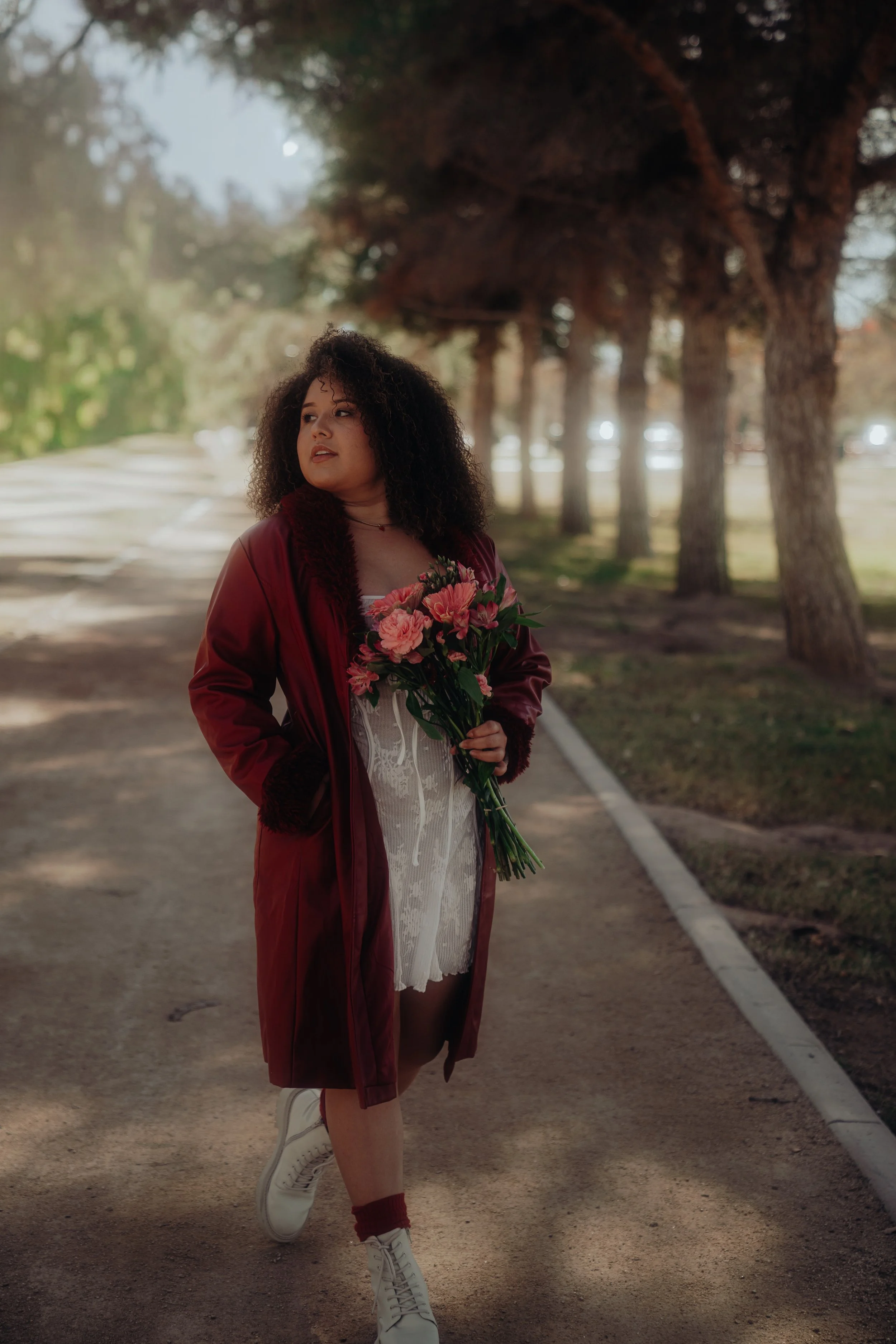 A young woman with curly hair wearing a red satin jacket, a white lace dress, and white sneakers. She is holding a bouquet of pink flowers and standing on a dirt path in a park with tall trees and dappled sunlight.