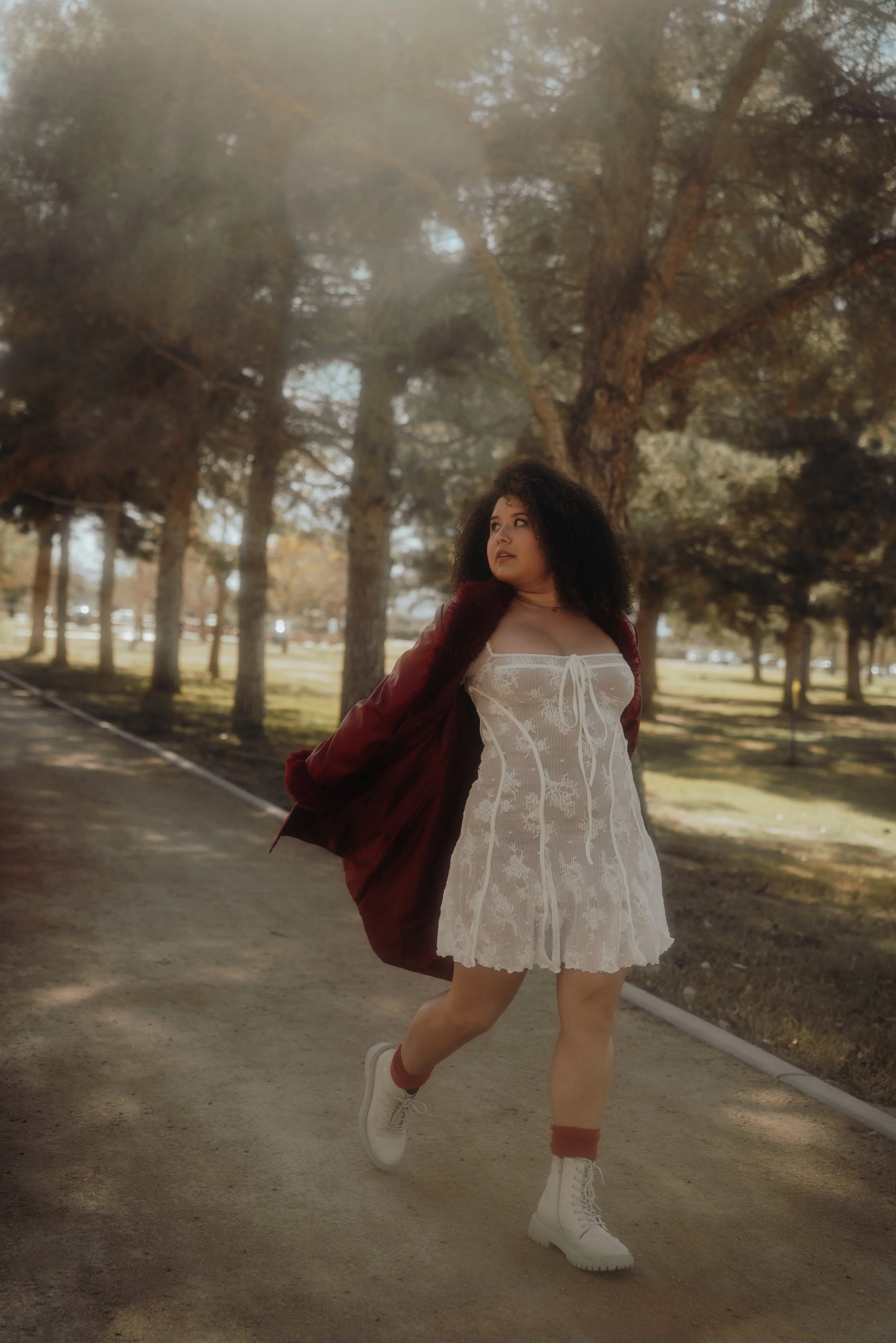 A woman with curly dark hair is walking on a dirt path in a park with tall trees. She is wearing a white lace dress, a maroon jacket draped over her shoulders, white boots, and maroon socks.