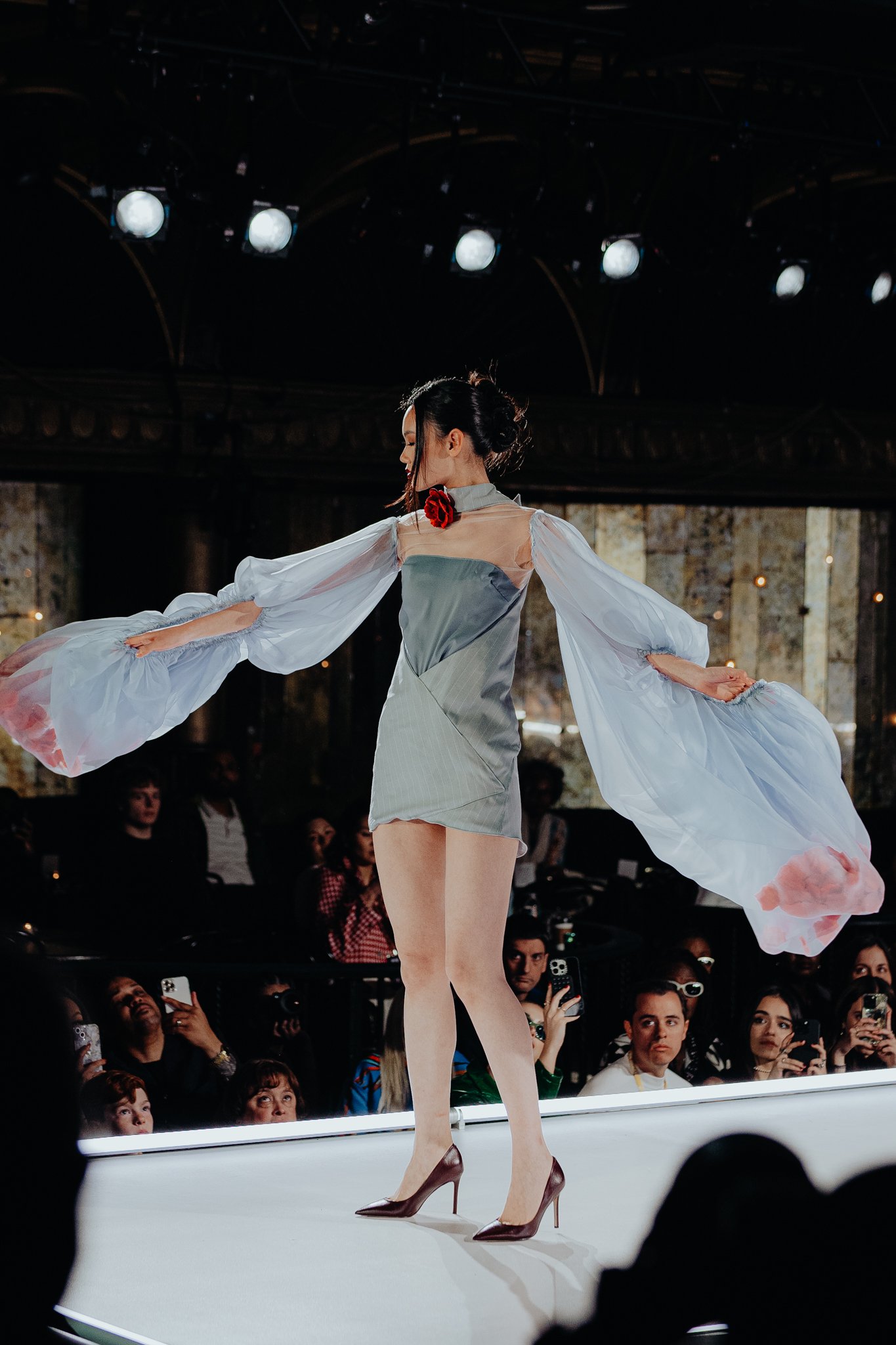 A woman walking on a runway at a fashion show, wearing a fashion-forward outfit with flowing, sheer sleeves, high heels, and a red flower accessory on her neck. Audience members are visible in the background, watching and taking pictures.