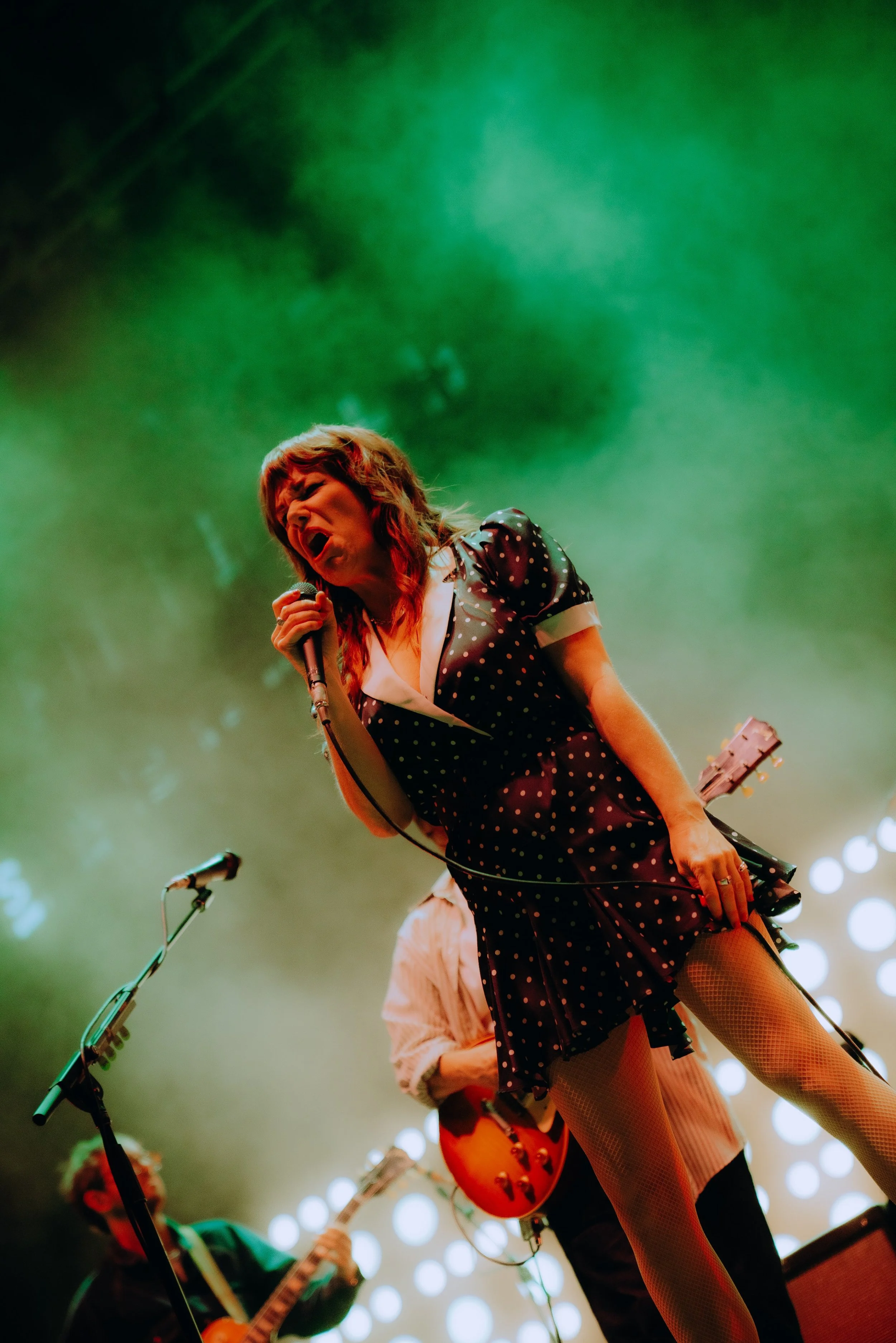 A female singer in a black polka dot dress passionately singing on stage, with a guitarist to her left playing an electric guitar, both illuminated by stage lights with a dotted circular pattern in the background.