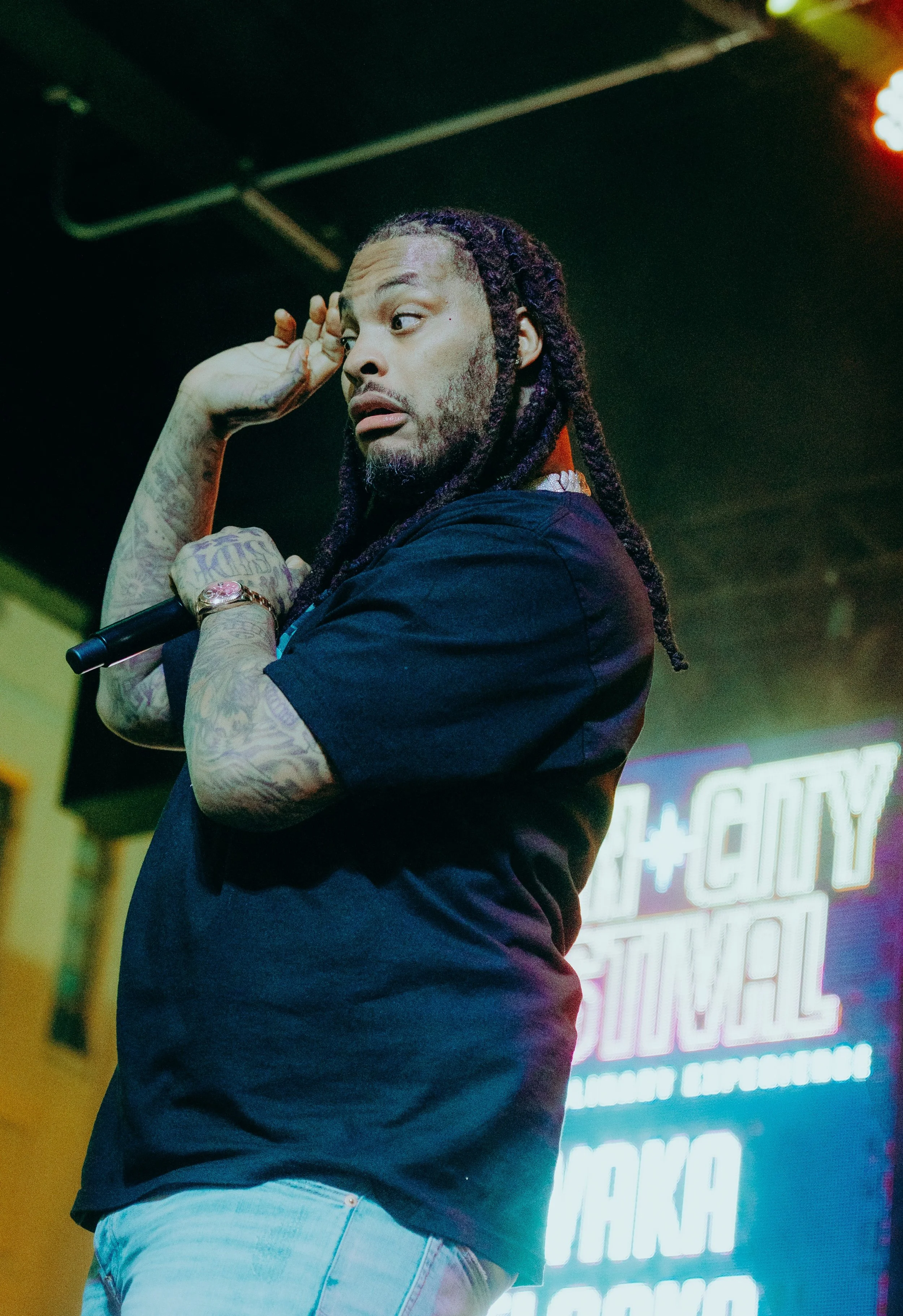A man with long dreadlocks and facial hair stands in a dimly lit space, holding a microphone, with neon signs in the background.