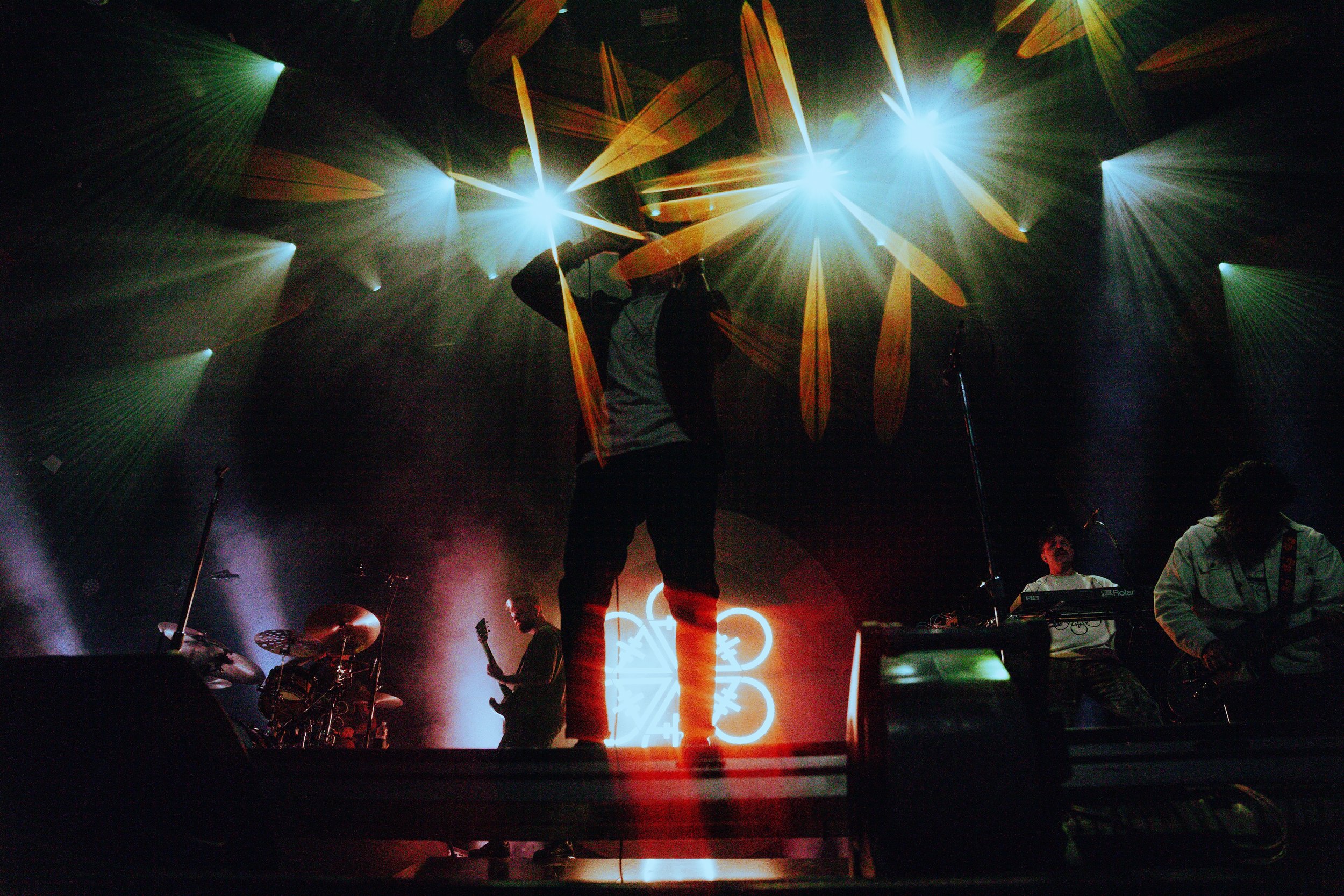 A concert scene featuring a band playing on stage with colorful lights and laser effects. The lead performer is in the center, raising their hand, surrounded by musicians and stage equipment.