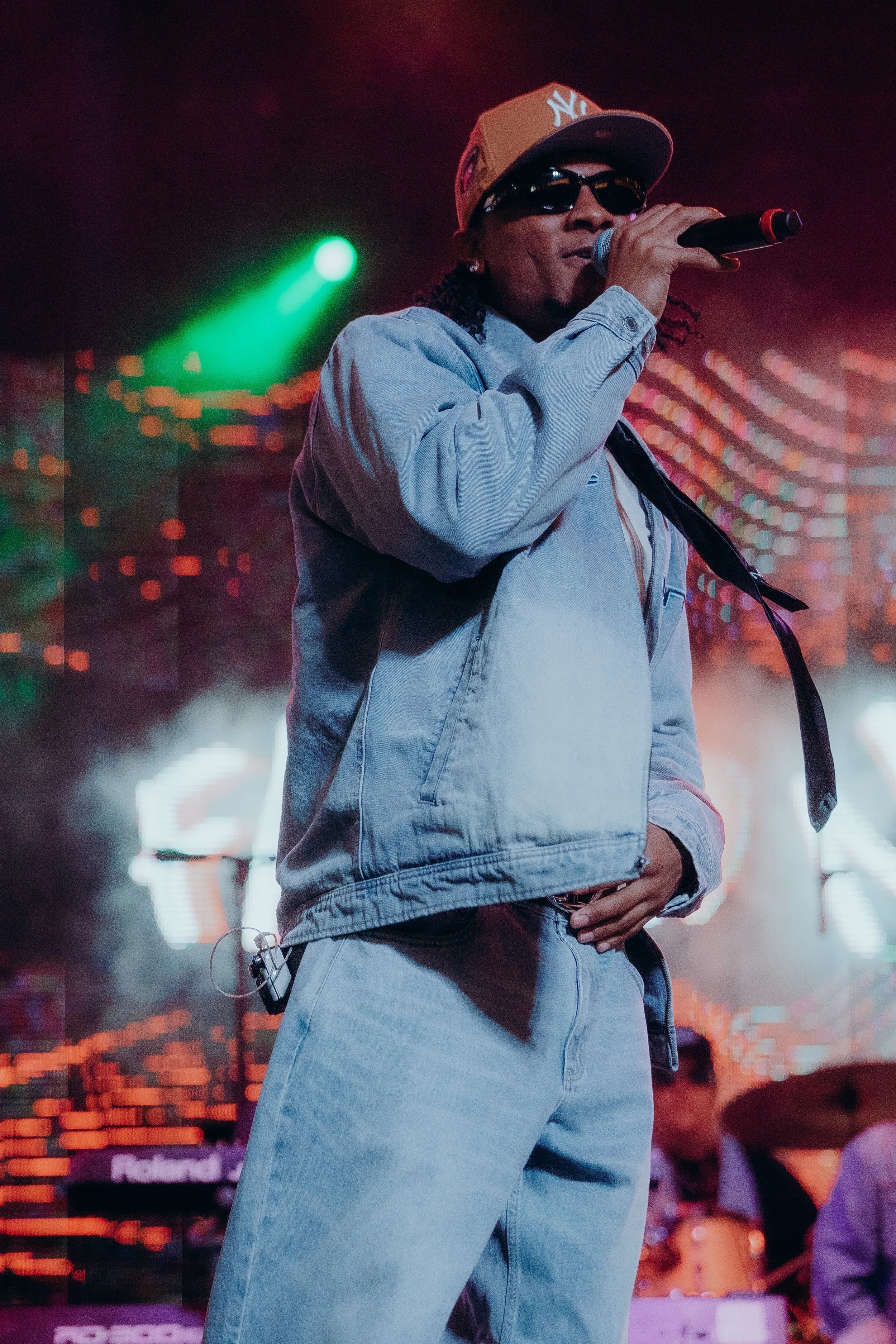 A man wearing a gray New York Yankees baseball cap, black sunglasses, and a light denim jacket, singing into a microphone on stage with colorful lights and a blurred background of a music venue.