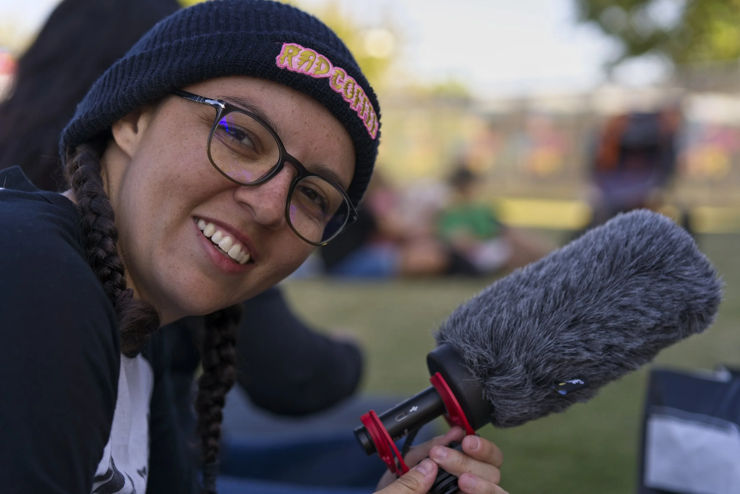 A young woman with glasses and braided hair wearing a knit beanie with the word "REDCORE" patch smiling while holding a camera with a large microphone. She is outdoors with blurred trees and people in the background.