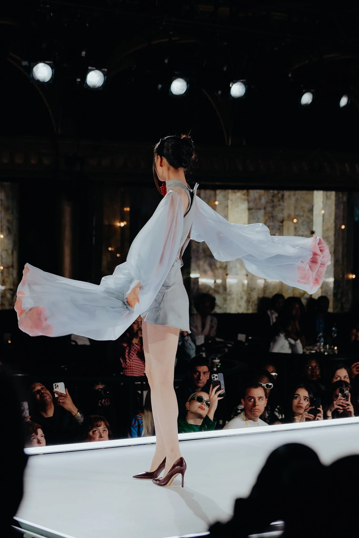 A woman is performing on stage in front of an audience, wearing a flowing dress and high heels. The audience is watching and taking photos.