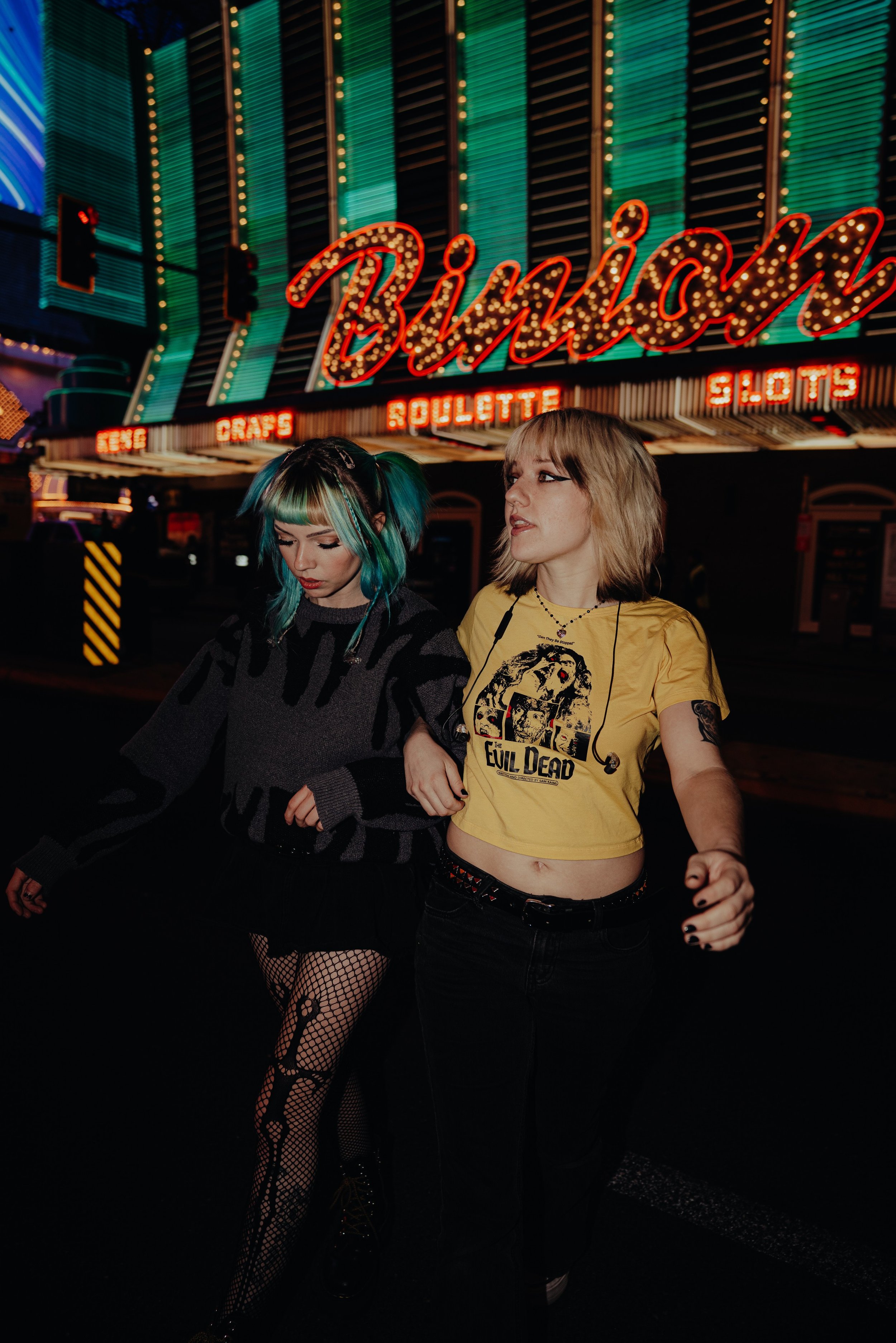 Two young women walking in front of a brightly lit Las Vegas casino sign. One has colorful hair and fishnet stockings, the other has blonde hair and wears a yellow T-shirt with a graphic design.