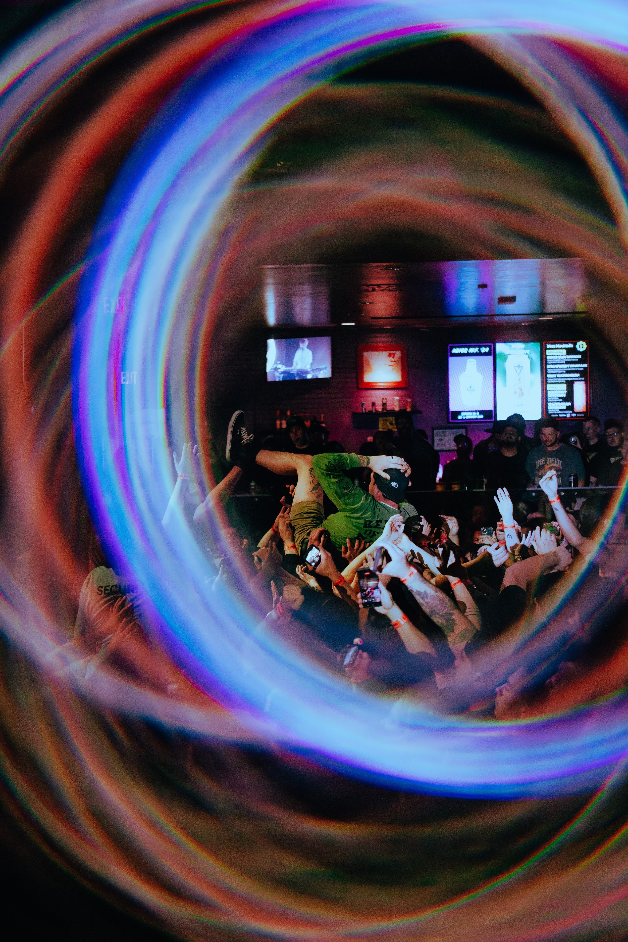 People crowd a concert venue, seen through a spiral-shaped mirror with colorful, swirling patterns, with a stage and screens visible in the background.