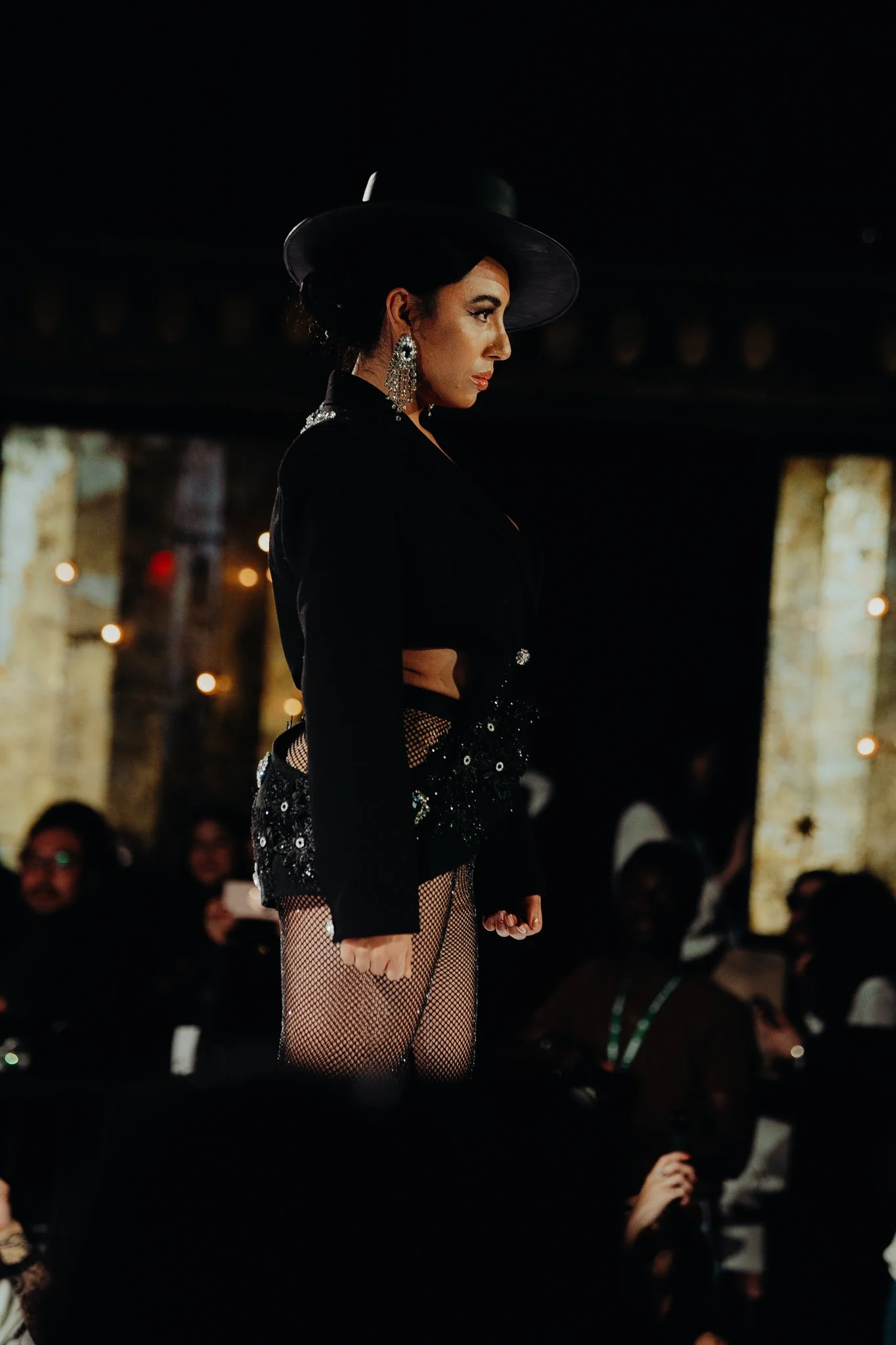 A woman performing on stage wearing a black wide-brimmed hat, large chandelier earrings, a black top, fishnet stockings, and a sequined skirt, with an audience in the background.