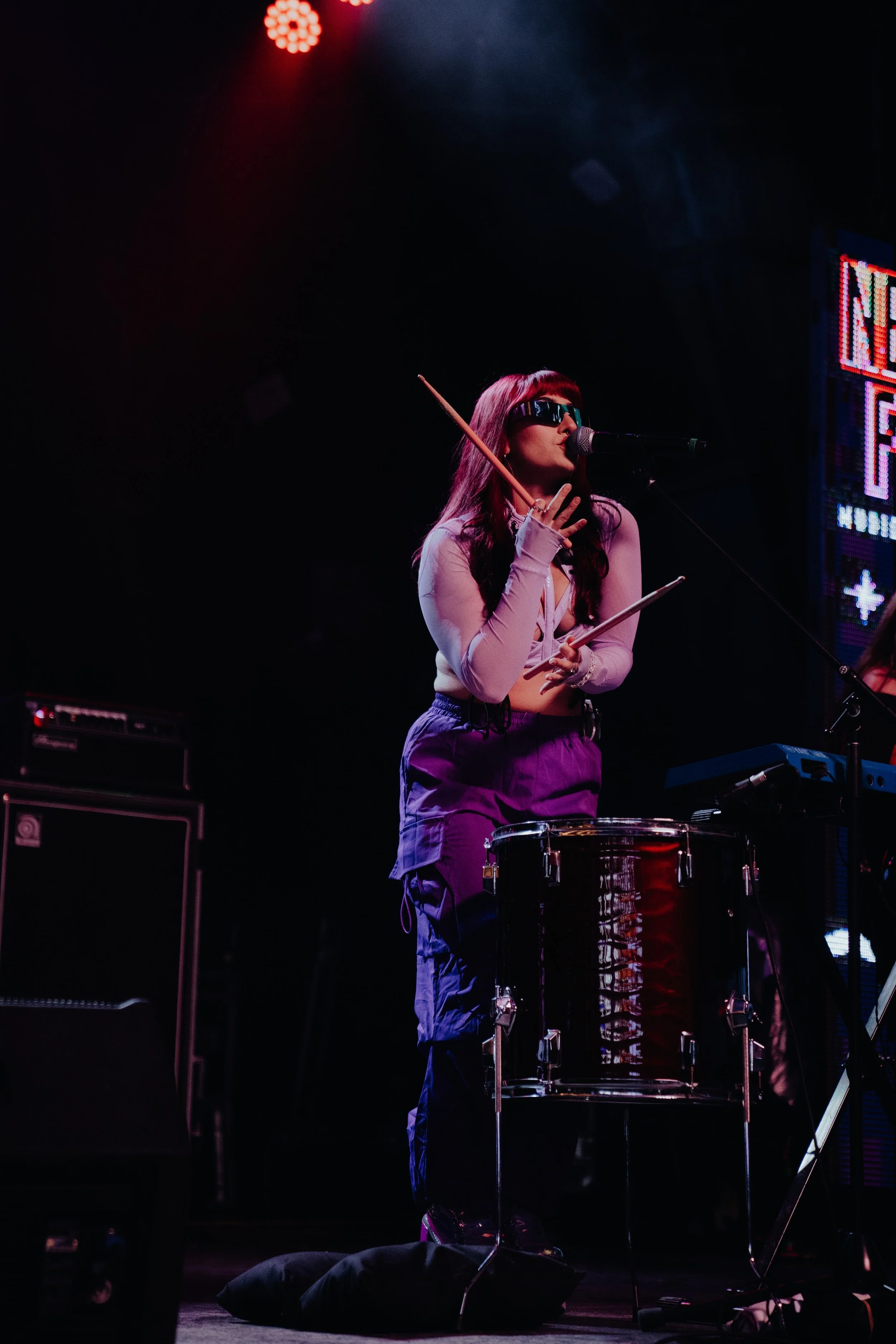 A woman wearing futuristic glasses is performing on stage, singing into a microphone while playing drums. She is dressed in a purple outfit with long sleeves and has long, dark hair. Stage equipment and LED screens with colorful lights are visible in