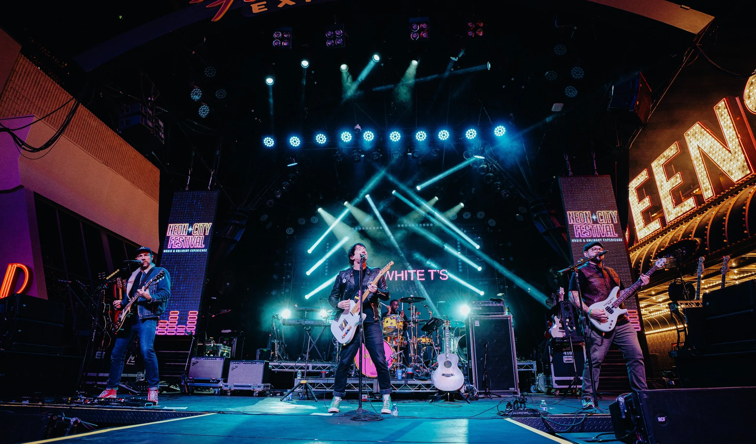 Band performing live on stage at Neon City Festival, with bright stage lights and festival signs visible.