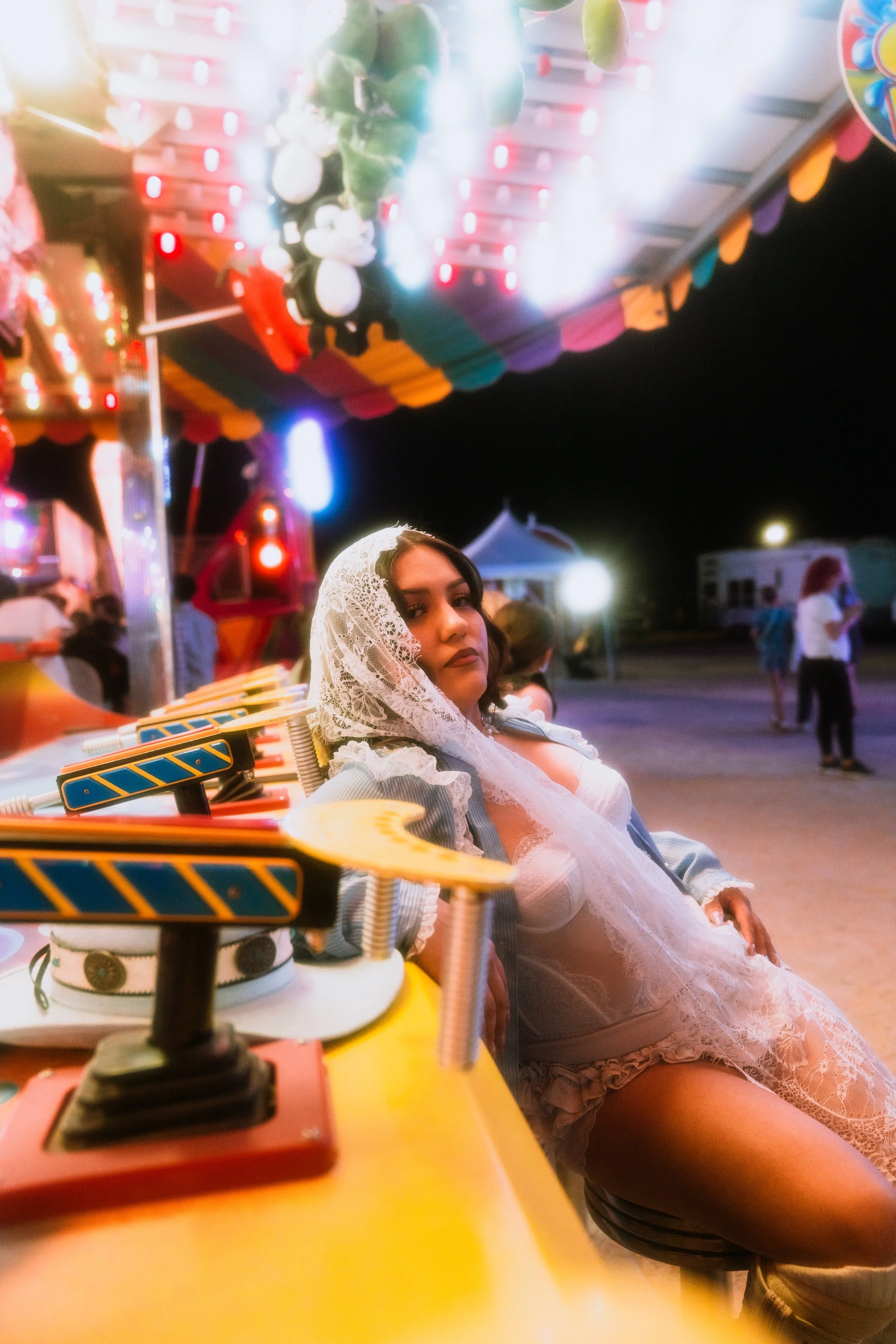 A woman with a lace veil sitting on a carnival ride at night, with brightly lit amusement rides and people in the background.