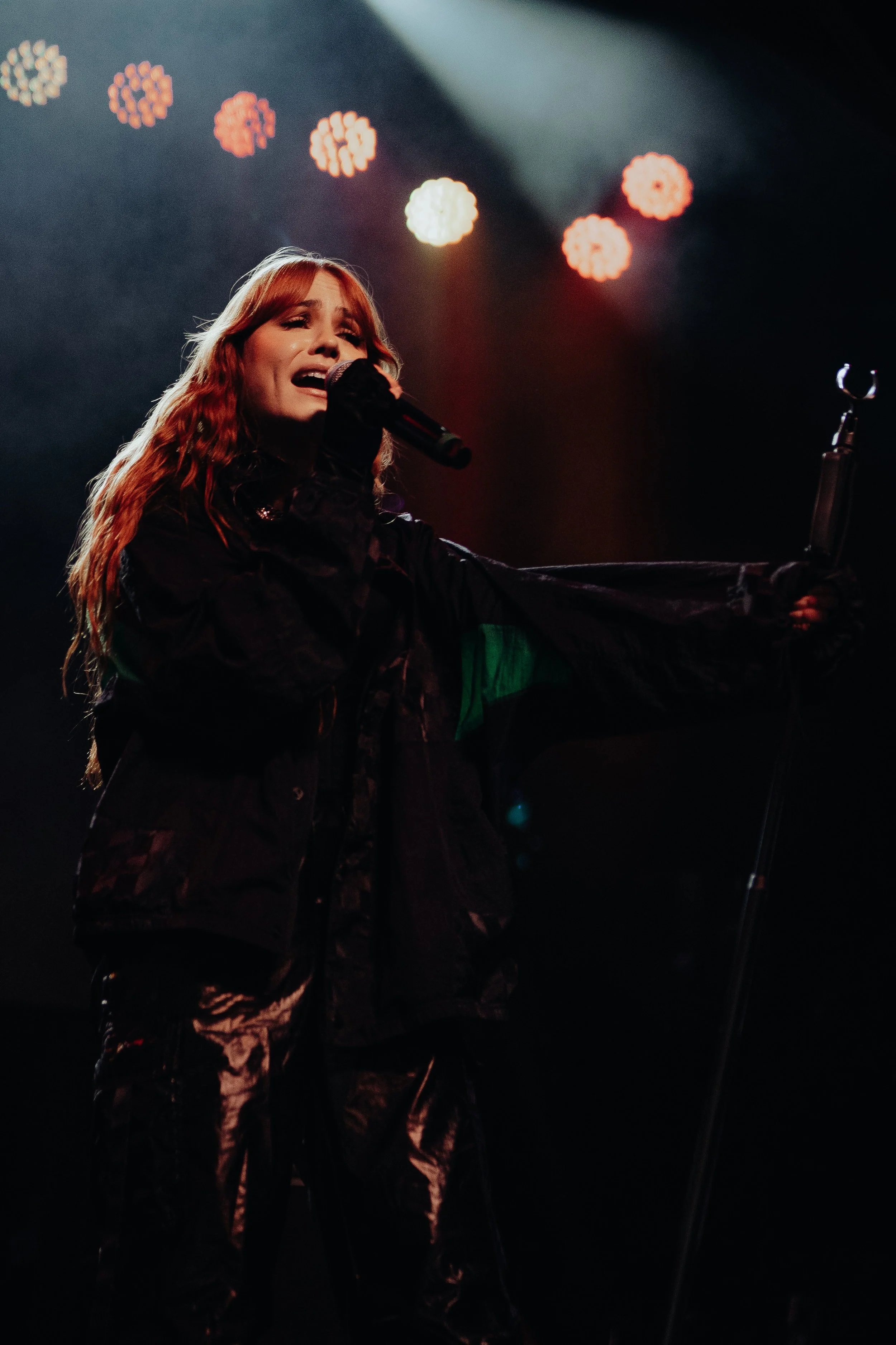 A female singer with long, wavy red hair performing on stage, holding a microphone with one hand, with colorful stage lights and a dark background.