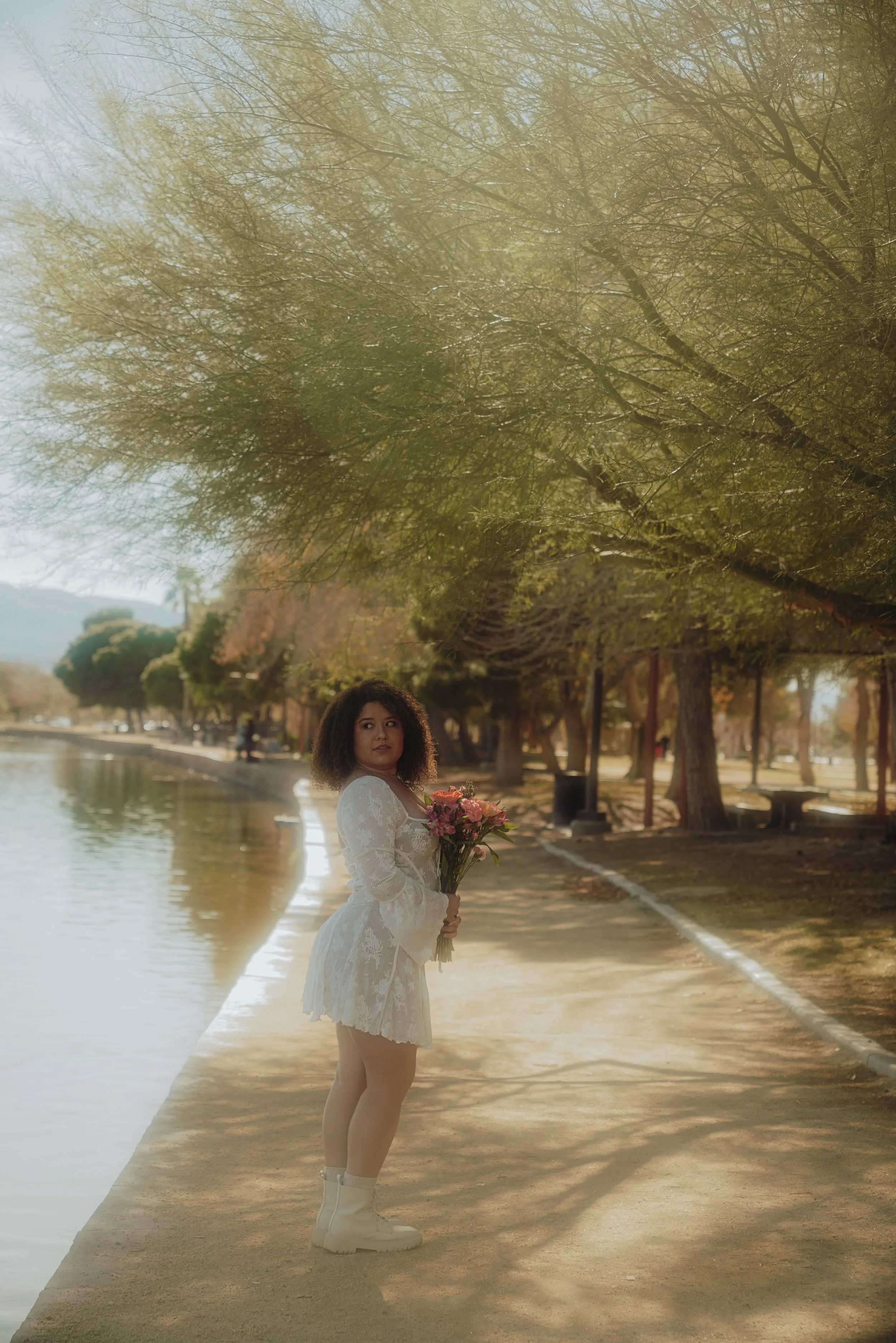 A woman with curly brown hair in a white lace dress holding a bouquet of pink and orange flowers, standing along a riverbank with trees and a sidewalk in the background during daytime.