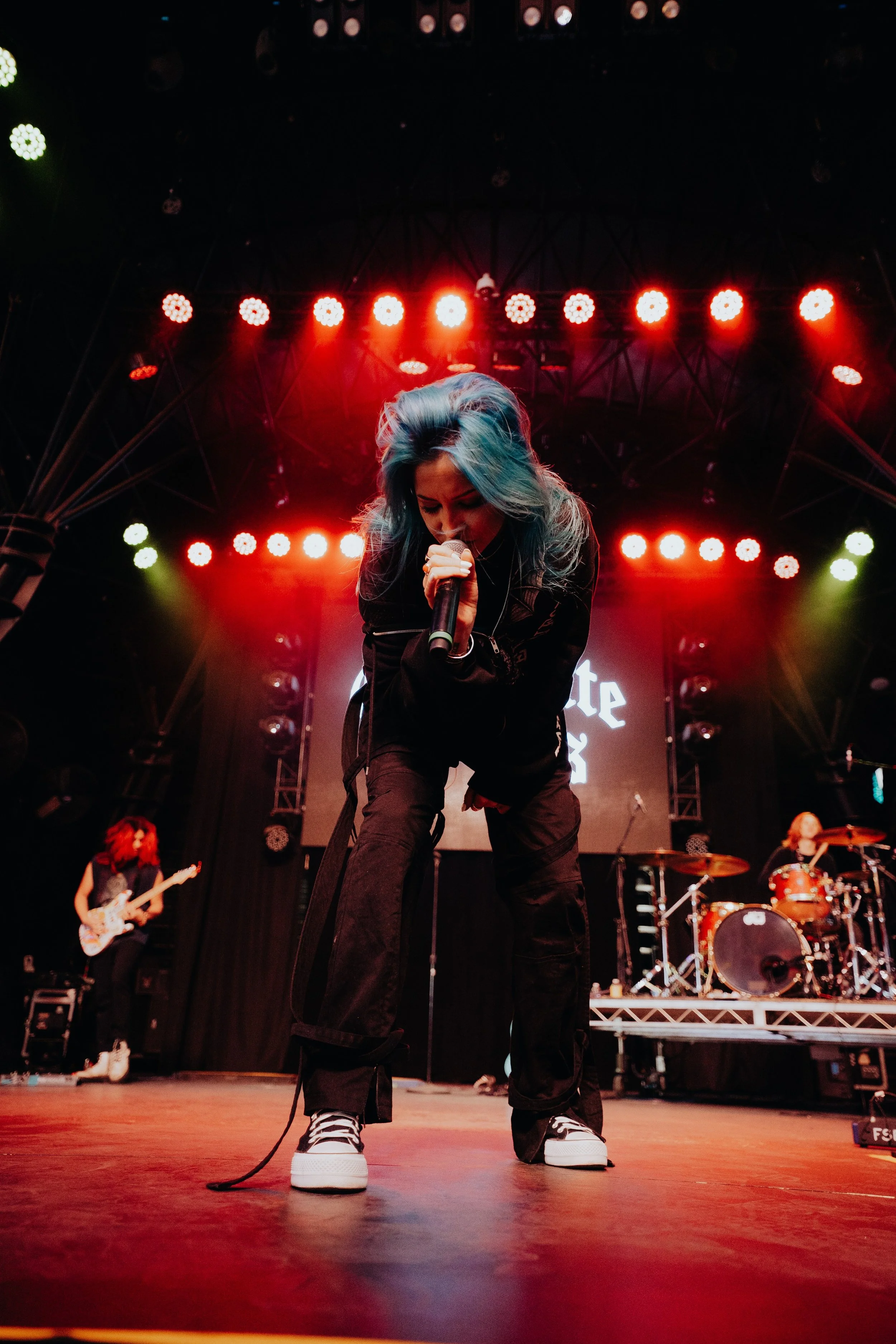 A female singer with blue hair performing on stage with musicians, red stage lights, and a digital backdrop.