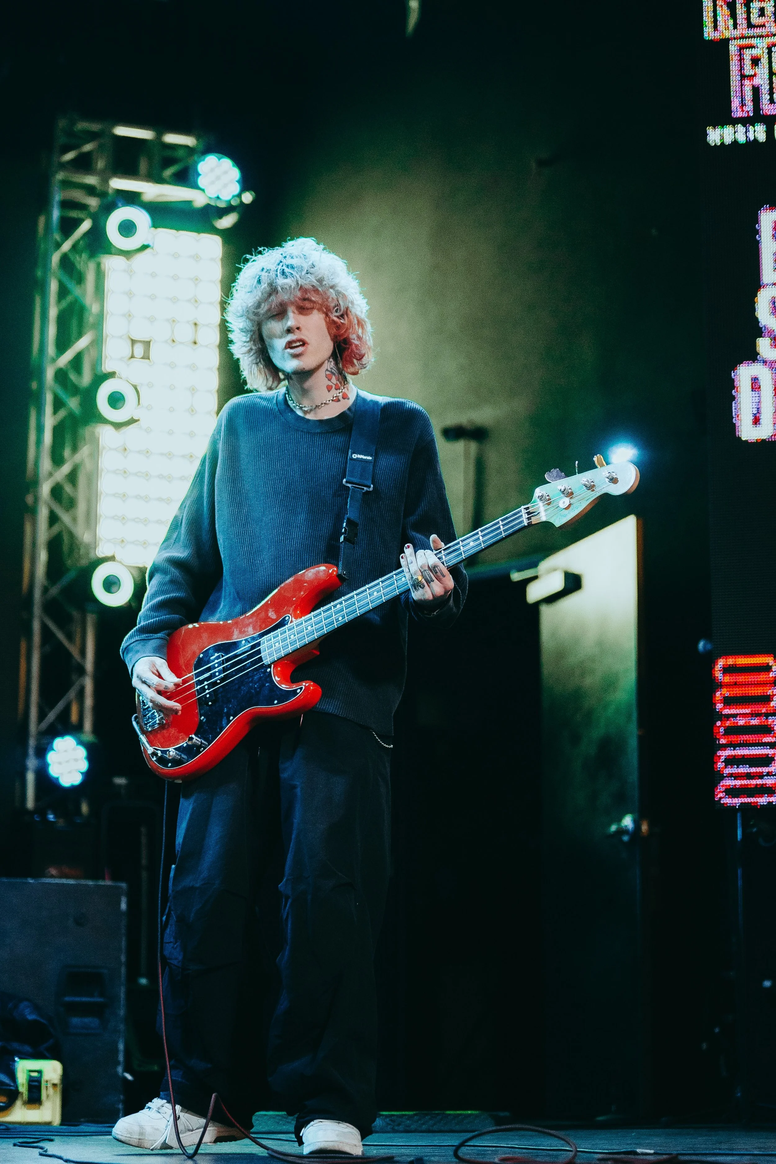 A young person with curly blonde hair playing an electric bass guitar on a stage with bright lights and colorful signage.