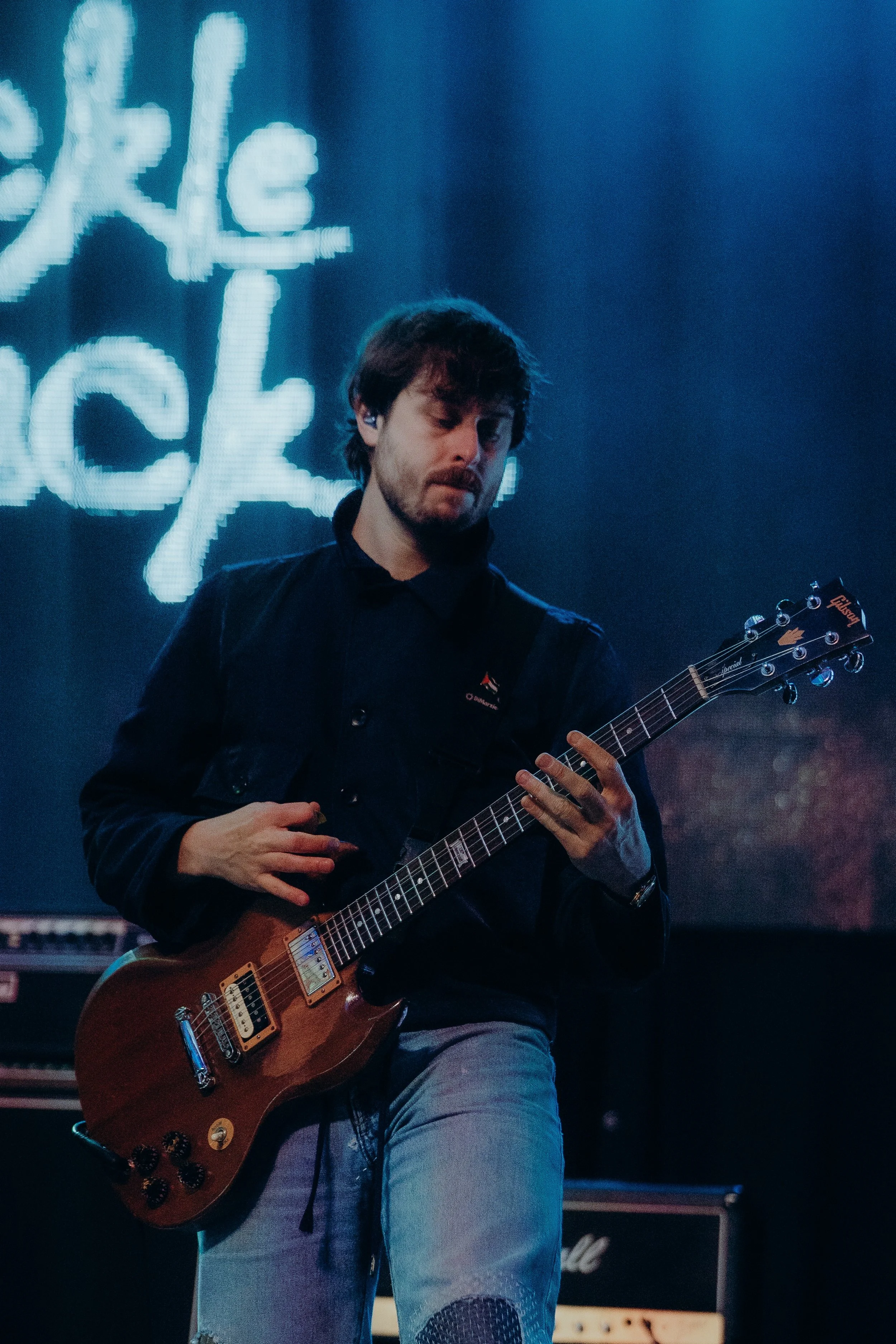 A man with brown hair and a beard playing an electric guitar on stage, wearing a black jacket and light-colored jeans, with a lit backdrop behind him.