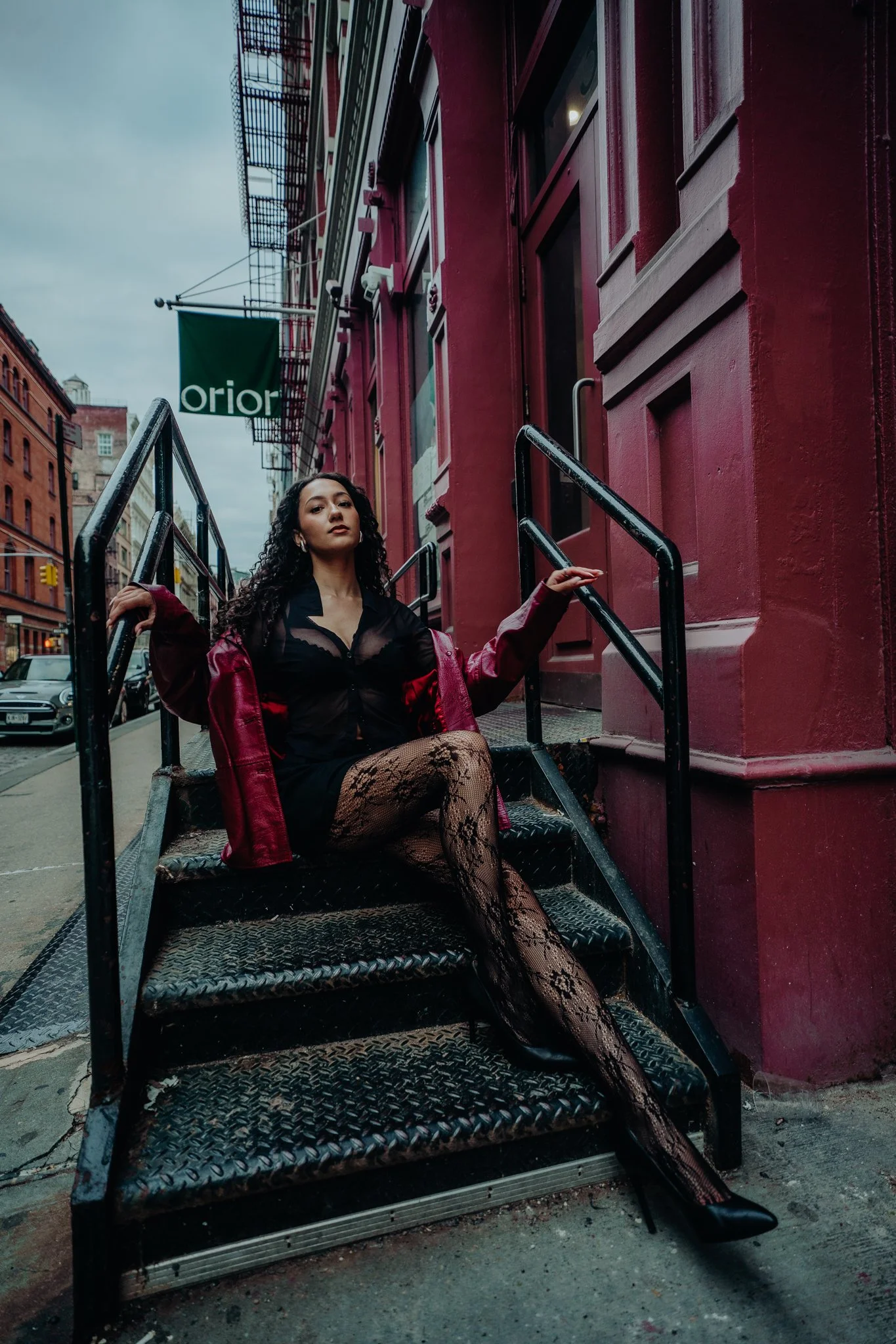 A woman with curly dark hair sitting on black metal stairs outside of a red brick building. She is wearing a sheer black dress, fishnet tights, black boots, and an unzipped burgundy jacket. The building features large windows and a green sign that re