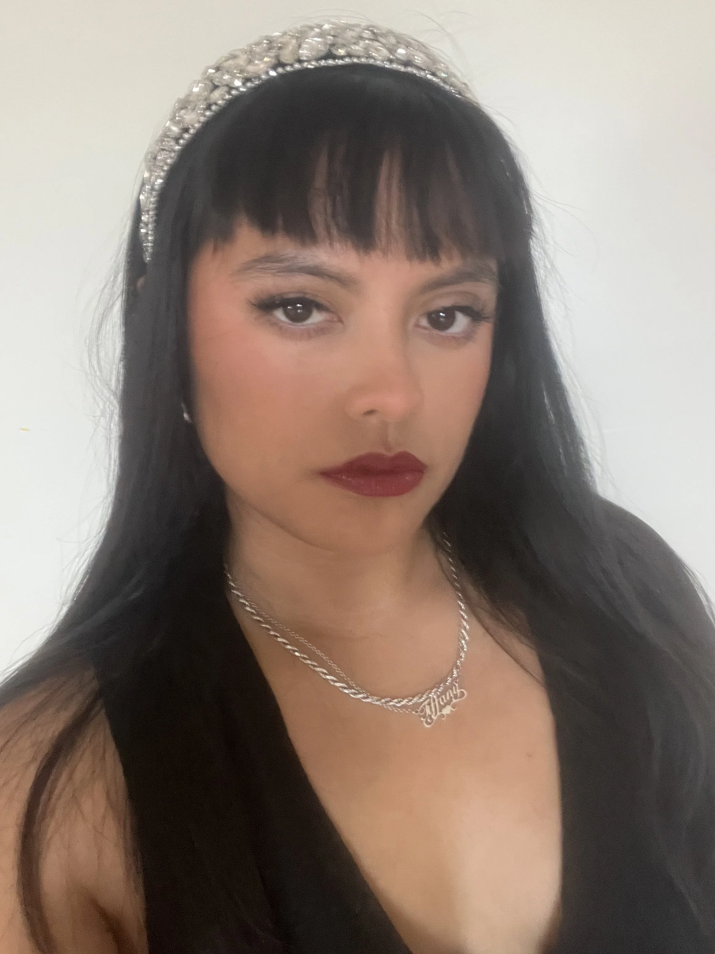 A woman with black hair, dark eye makeup, deep red lipstick, wearing a silver necklace and a glittery headband, looking into the camera against a plain background.