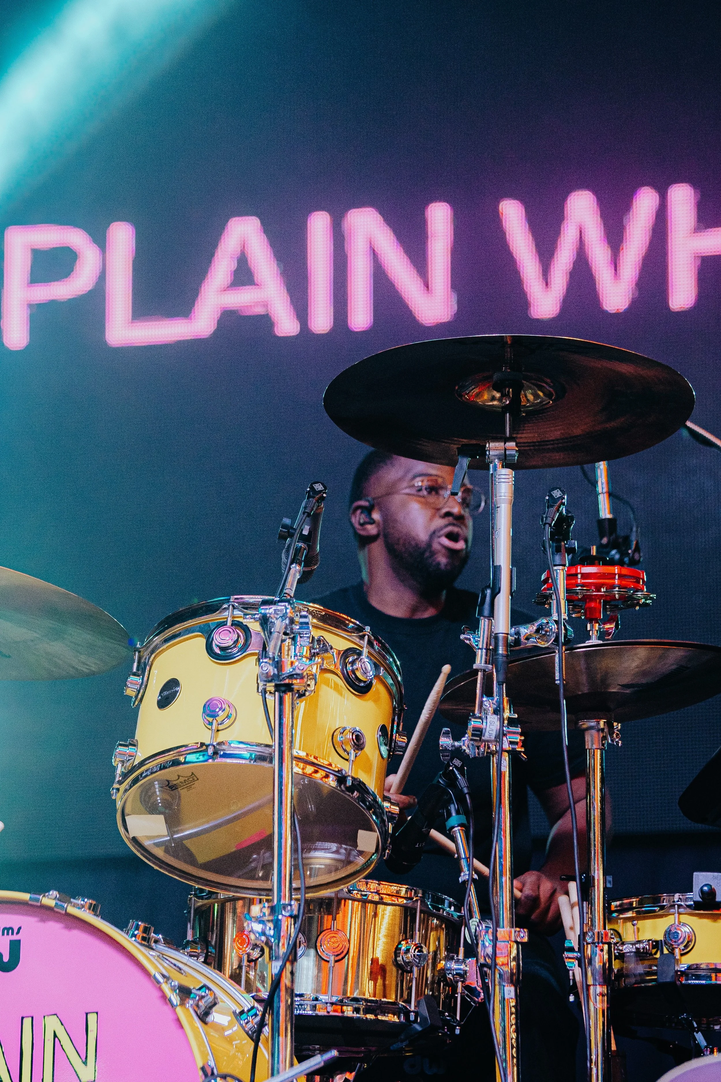 A man playing a yellow drum set with chrome hardware on stage, with a large pink neon sign in the background that reads 'PLAIN W.'