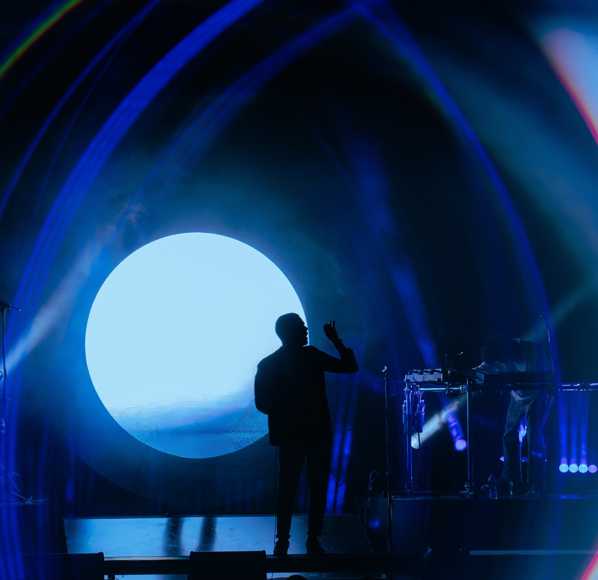 Silhouette of a person standing on stage with a microphone, in front of a large, bright blue circular light and blue curved light patterns, creating a dramatic, colorful scene.