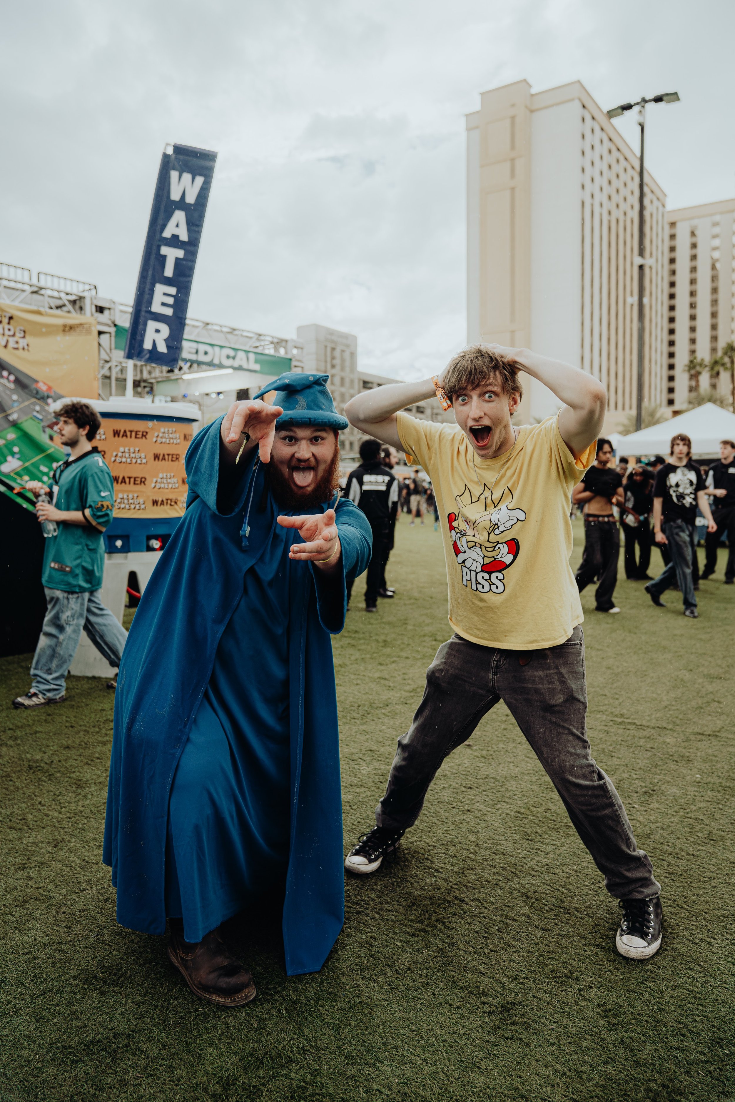Two young men are posing enthusiastically at an outdoor event, one dressed as a wizard with a blue cape and hat, pointing with a smile, while the other wears a yellow T-shirt with a graphic and 