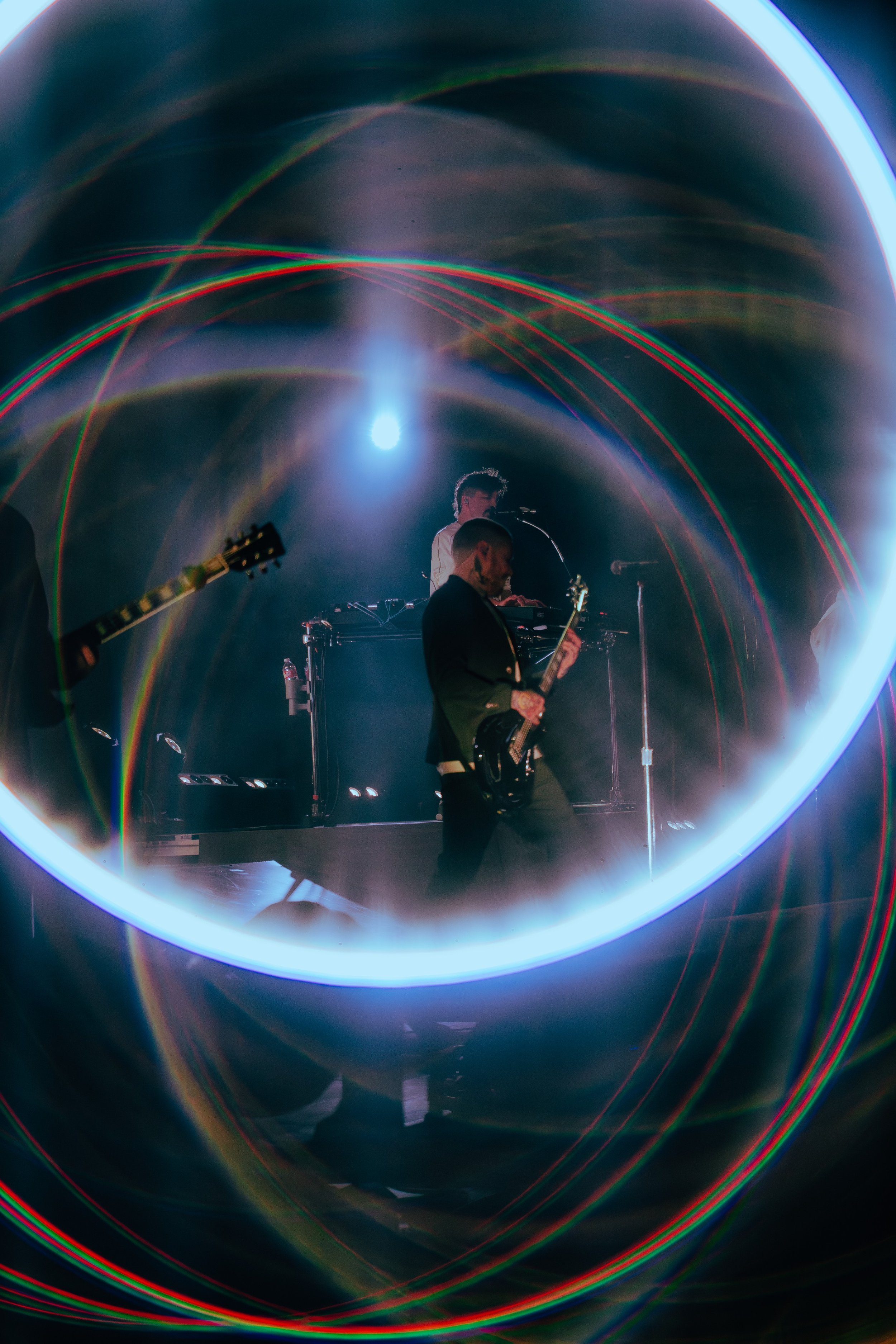 Musicians performing on stage with colorful laser lights creating circular patterns around them, including a person playing a guitar and another person using electronic equipment.