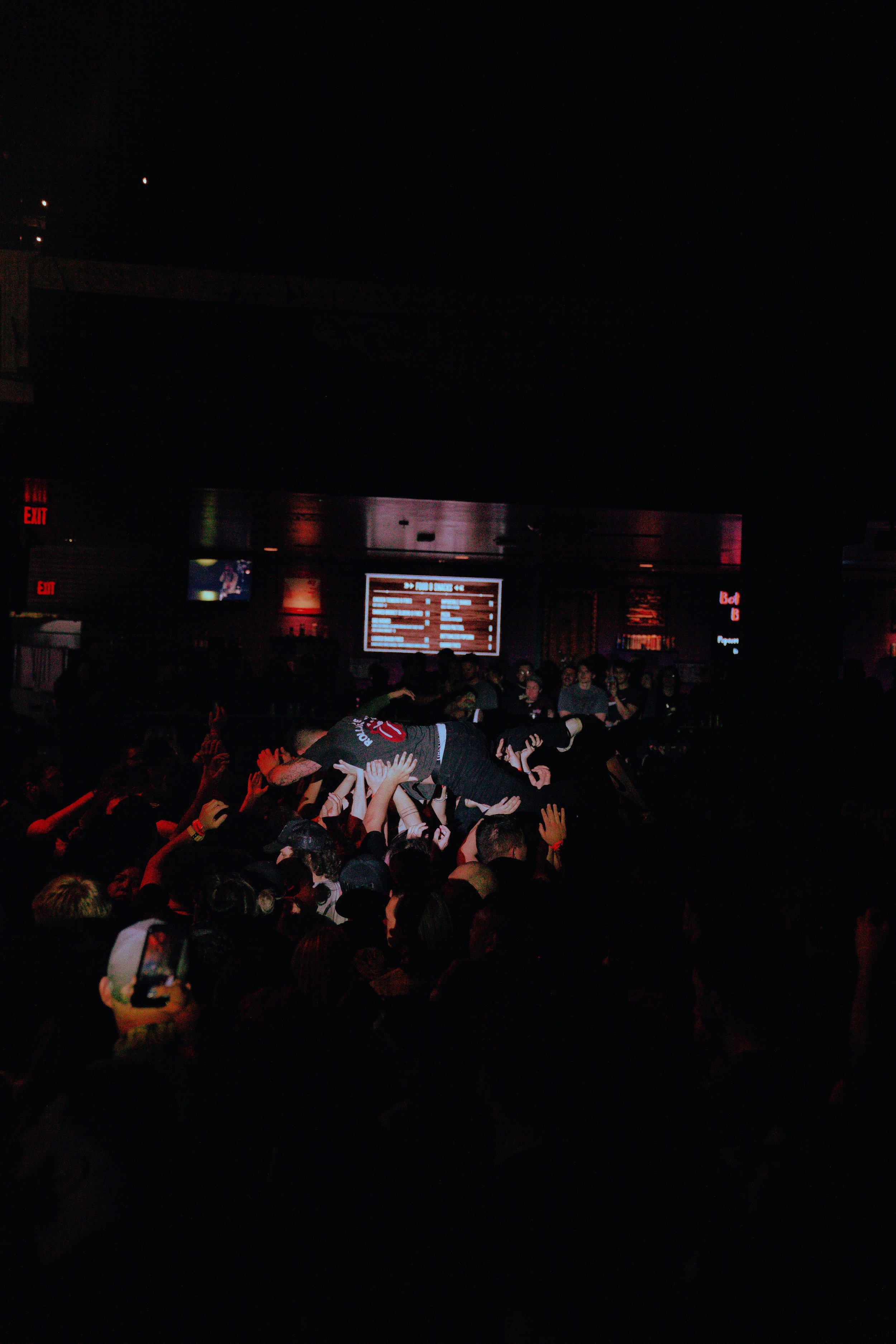 Crowd of people at a concert or nightclub with some individuals crowd surfing, dancing, and raising their hands, illuminated by stage lights and digital screens in the background.