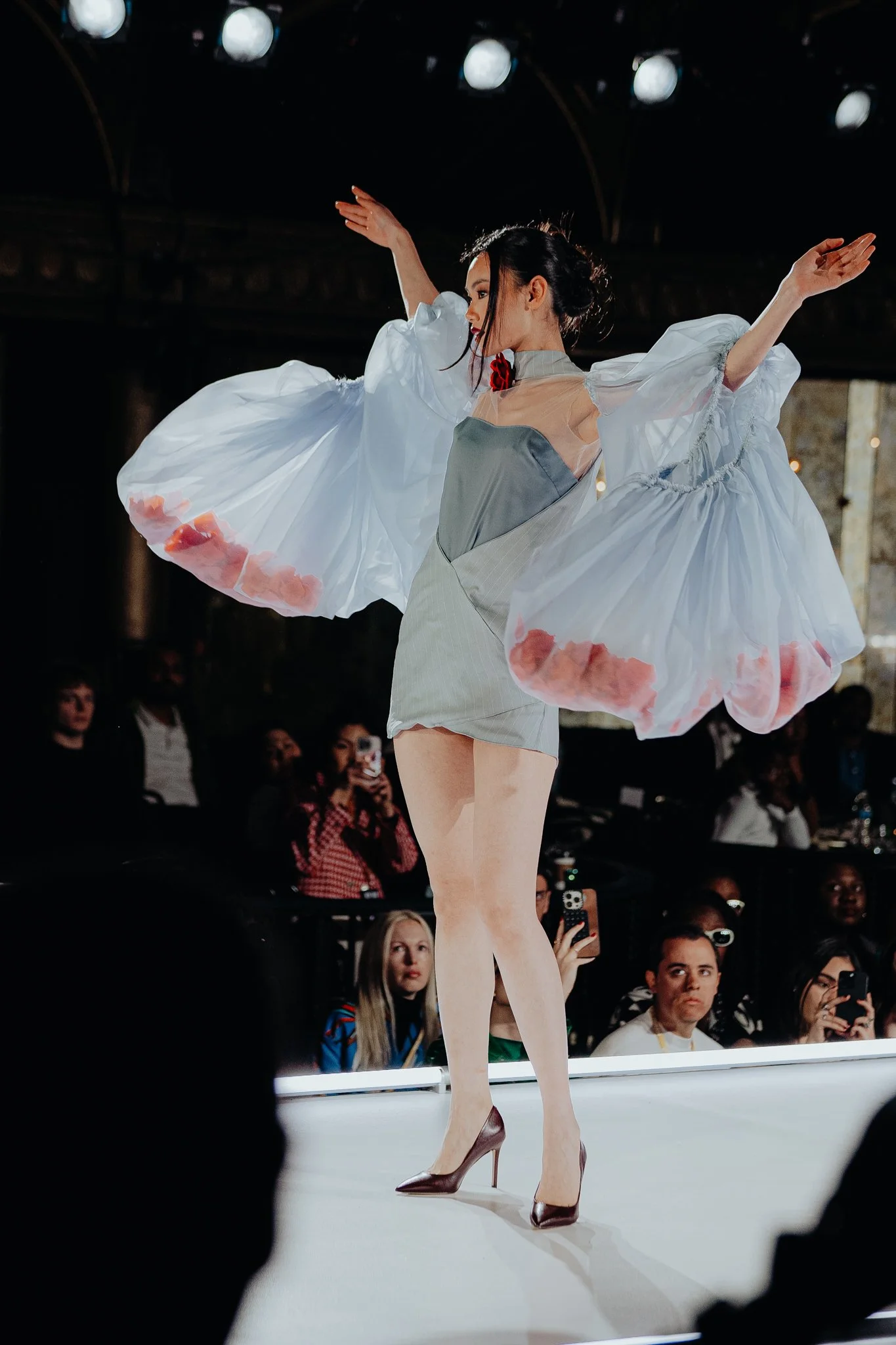 Fashion model on stage wearing beige high heels, sheer dress, and flowing skirts with pink floral accents, walking in front of an audience.