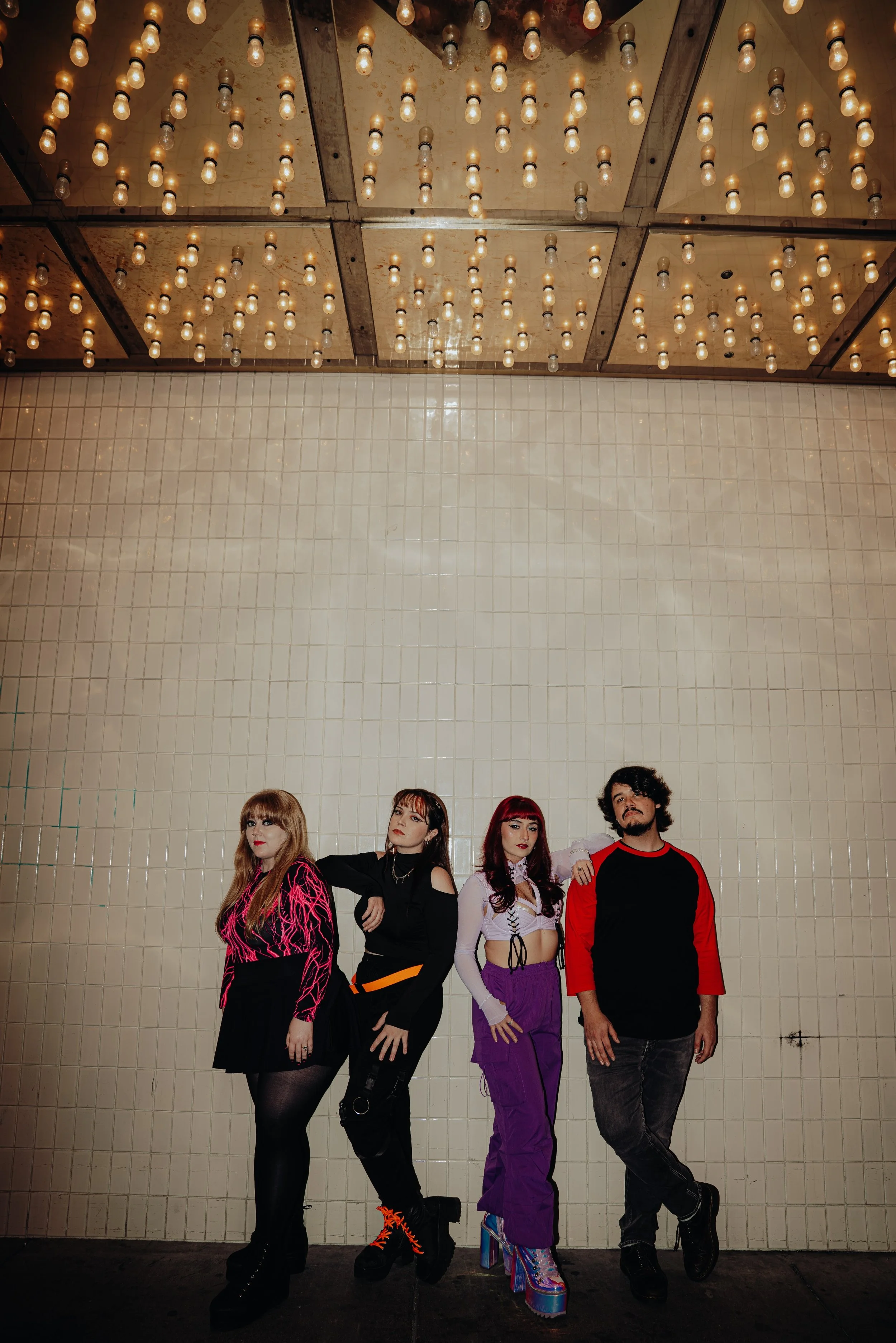 A group of five young adults posing in front of a tiled wall topped with a ceiling of exposed light bulbs.