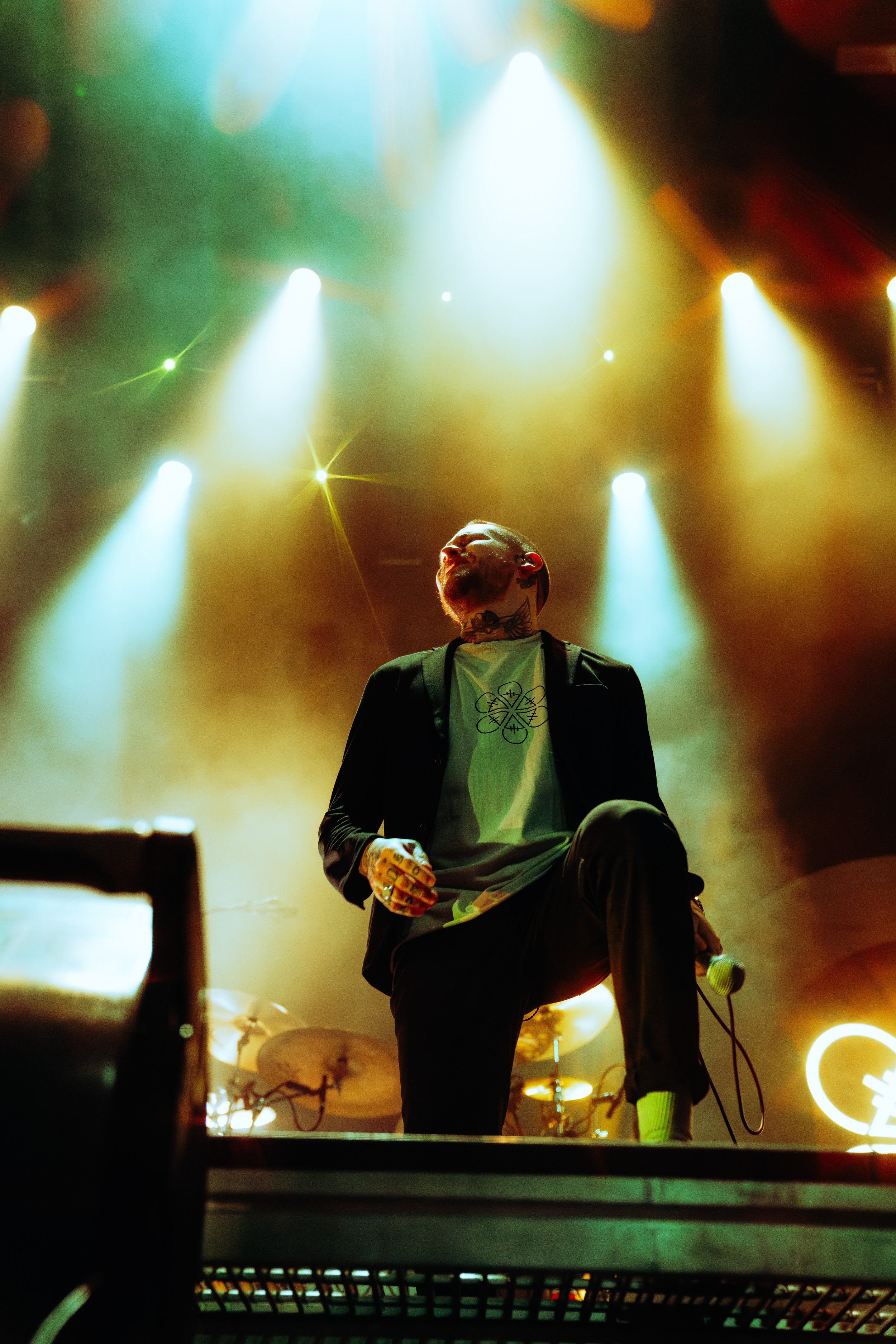 A performer with tattoos on their neck, holding a microphone, is on stage with colorful bright lights shining down, creating a concert atmosphere.
