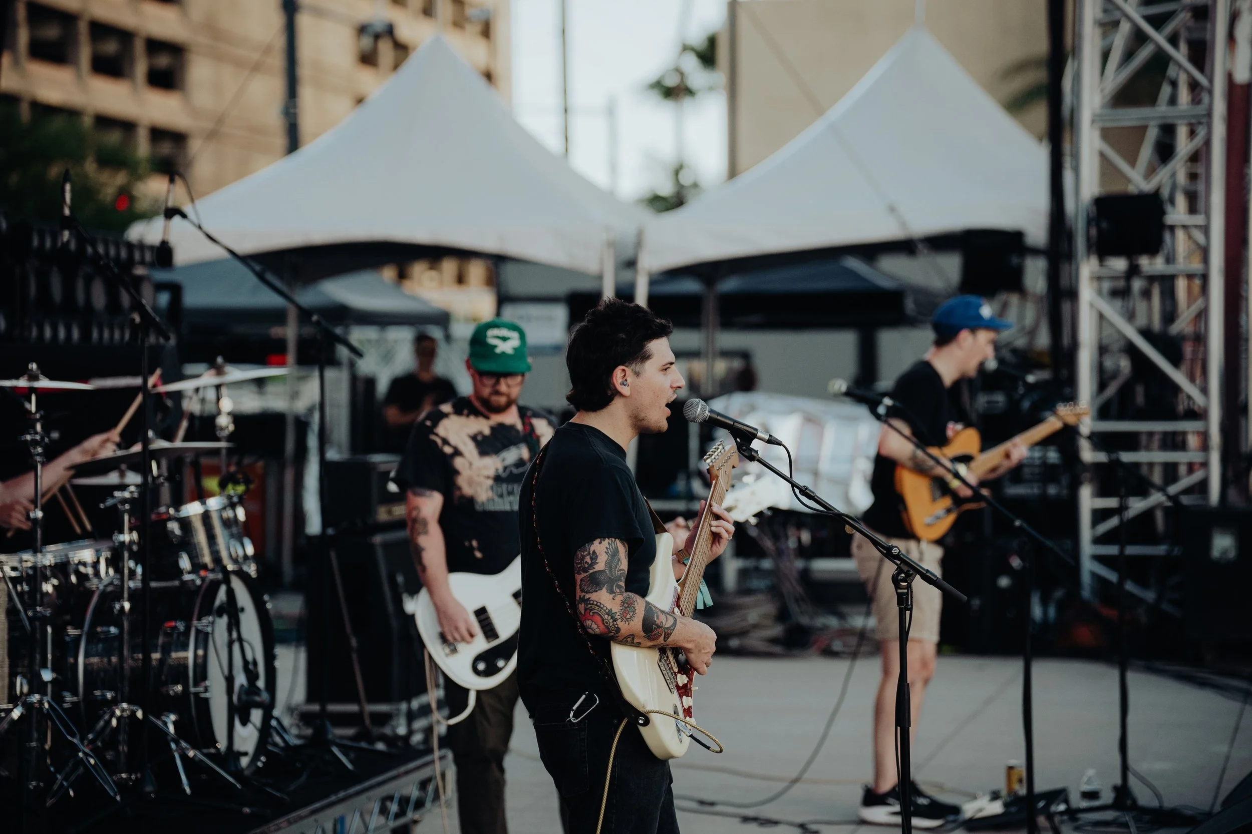 A band performs on an outdoor stage, with a lead singer playing an electric guitar and singing into a microphone, surrounded by fellow musicians with guitars and drums.