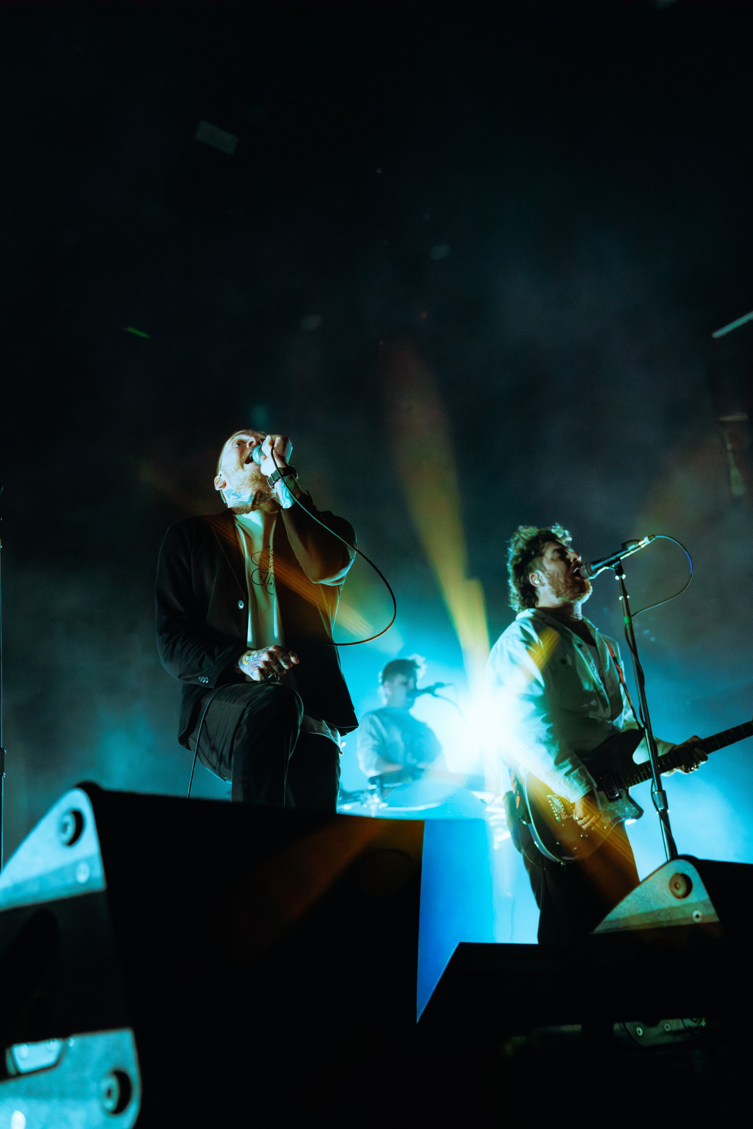 Live music performance with three male musicians on stage, one singing into a microphone and playing guitar, with colorful stage lights illuminating the scene.