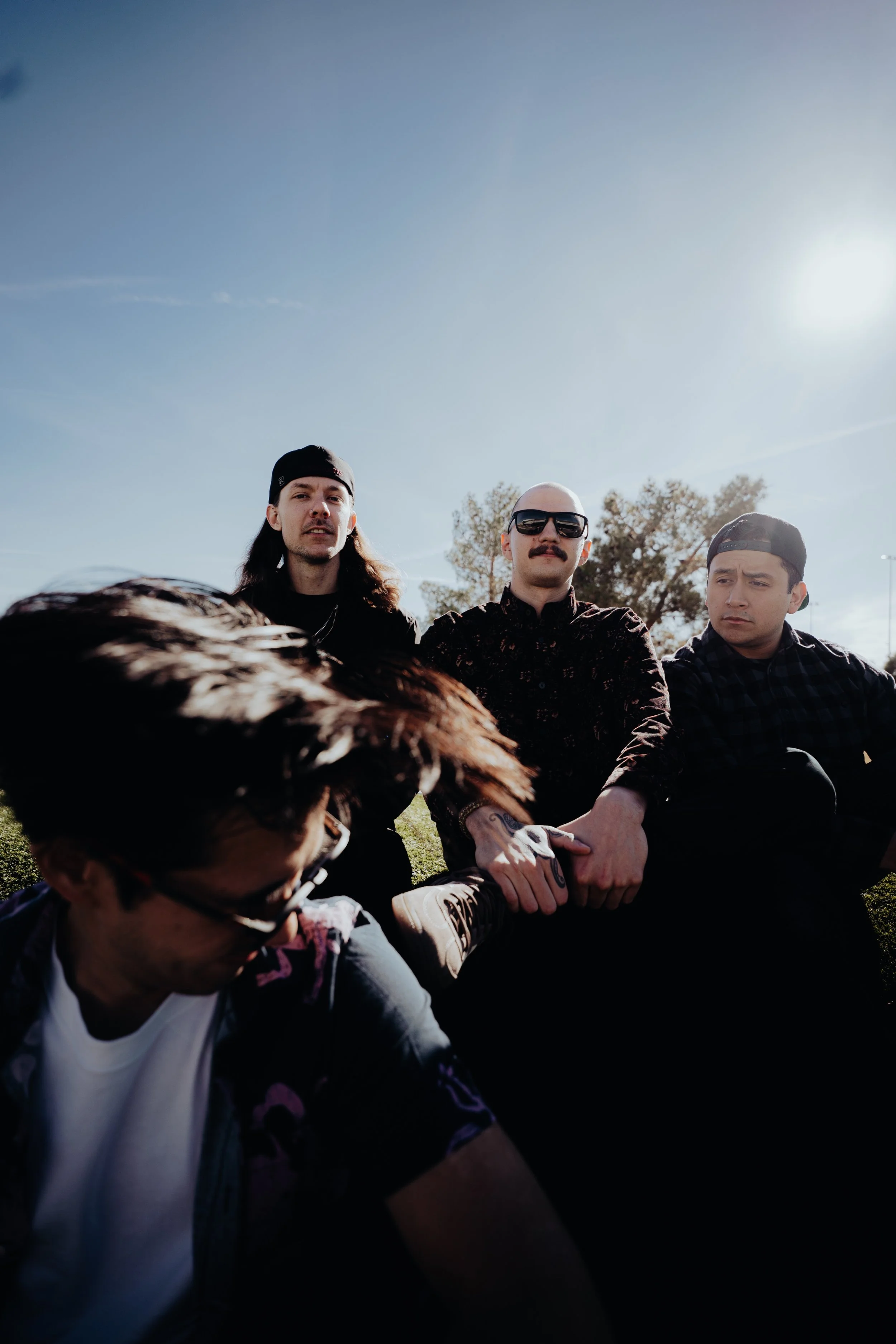 Three men sitting on the grass outdoors under a clear sky, with one person's head and hand in the foreground. The men are dressed casually, with one wearing sunglasses and the others wearing caps.
