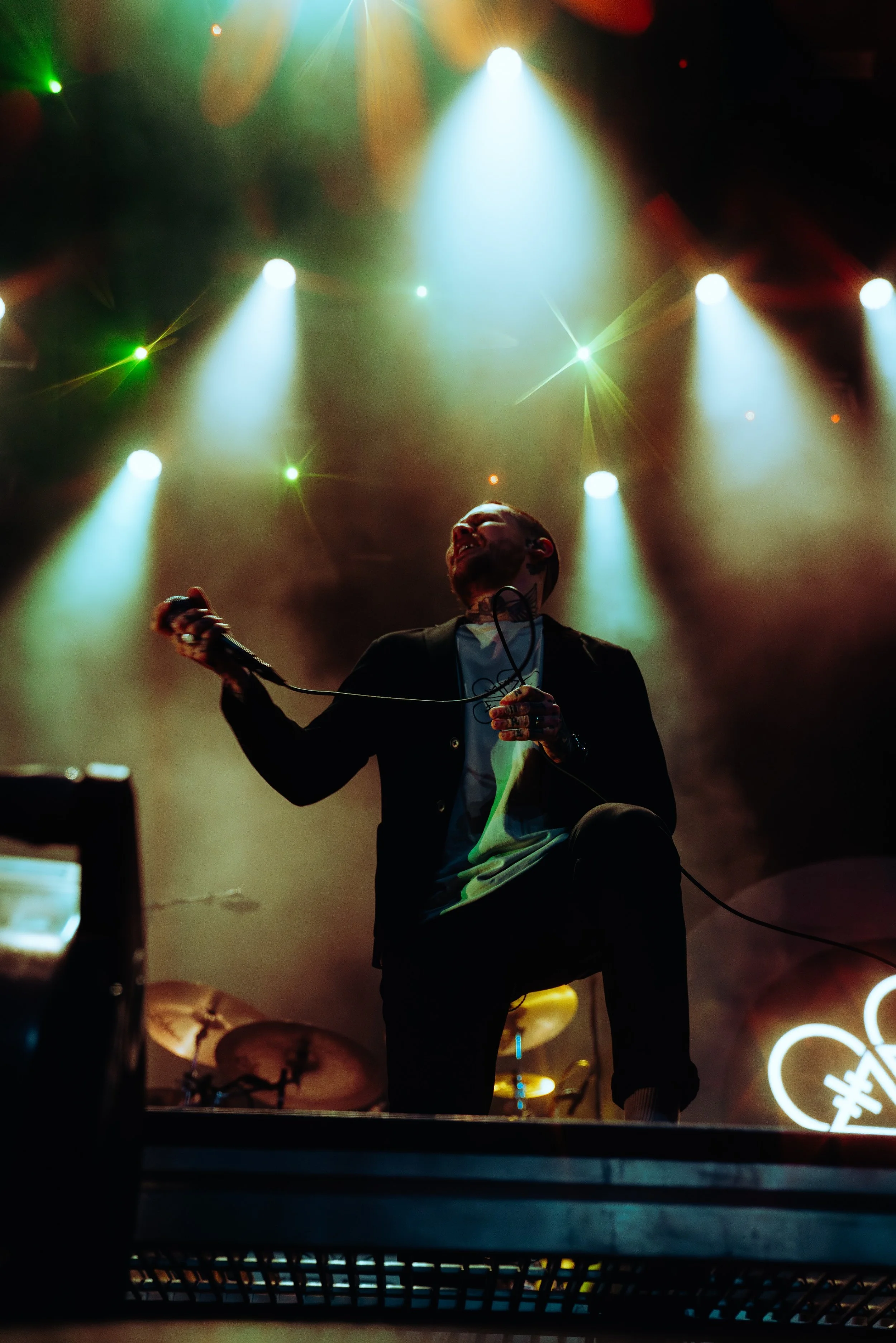 A singer performing on stage with colorful stage lights illuminating him, holding a microphone, with drums visible in the background.
