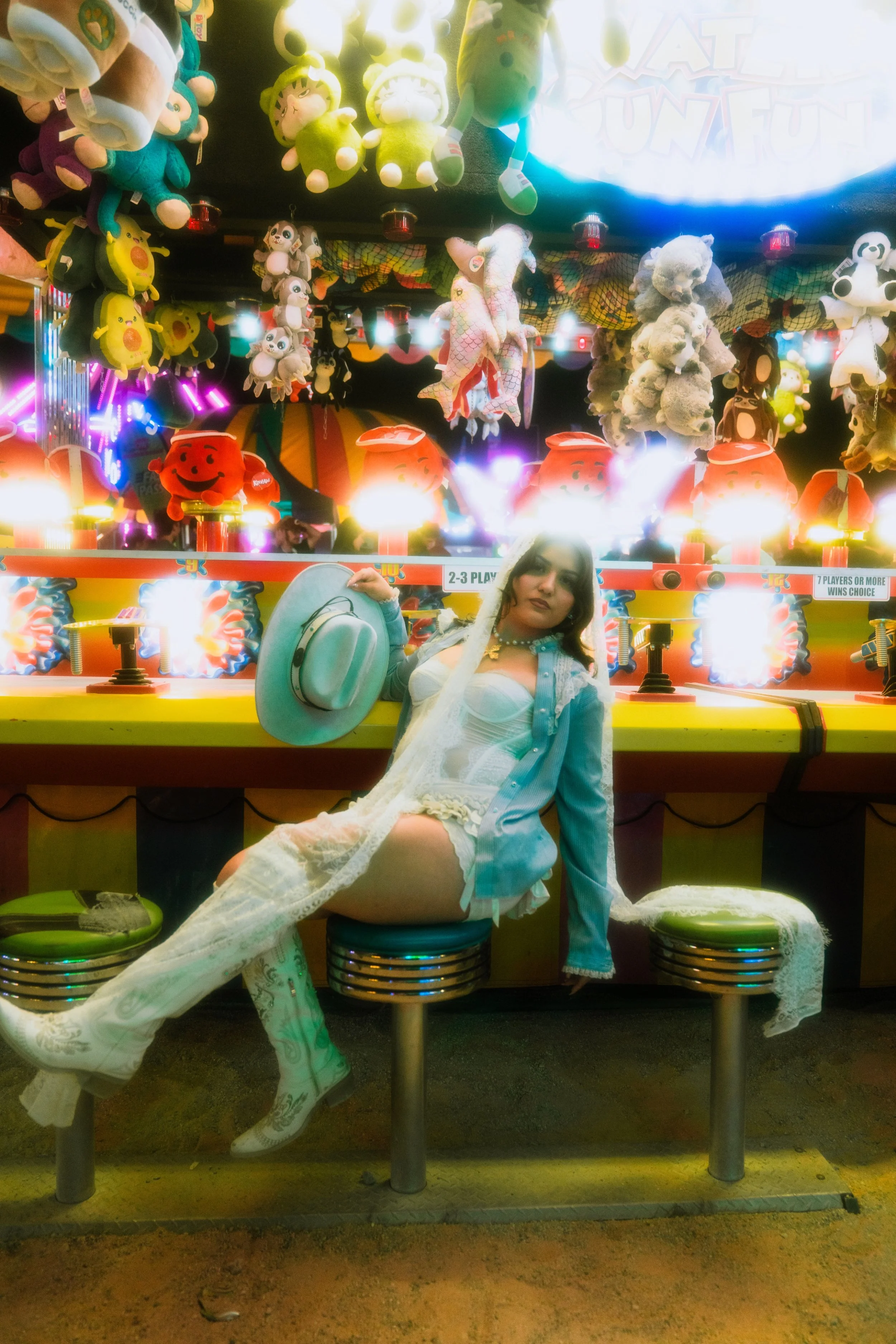 A young woman in a white lace outfit and cowboy hat sitting on a green stool in front of a carnival game with plush toys hanging in the background. The toys include various animals and characters.