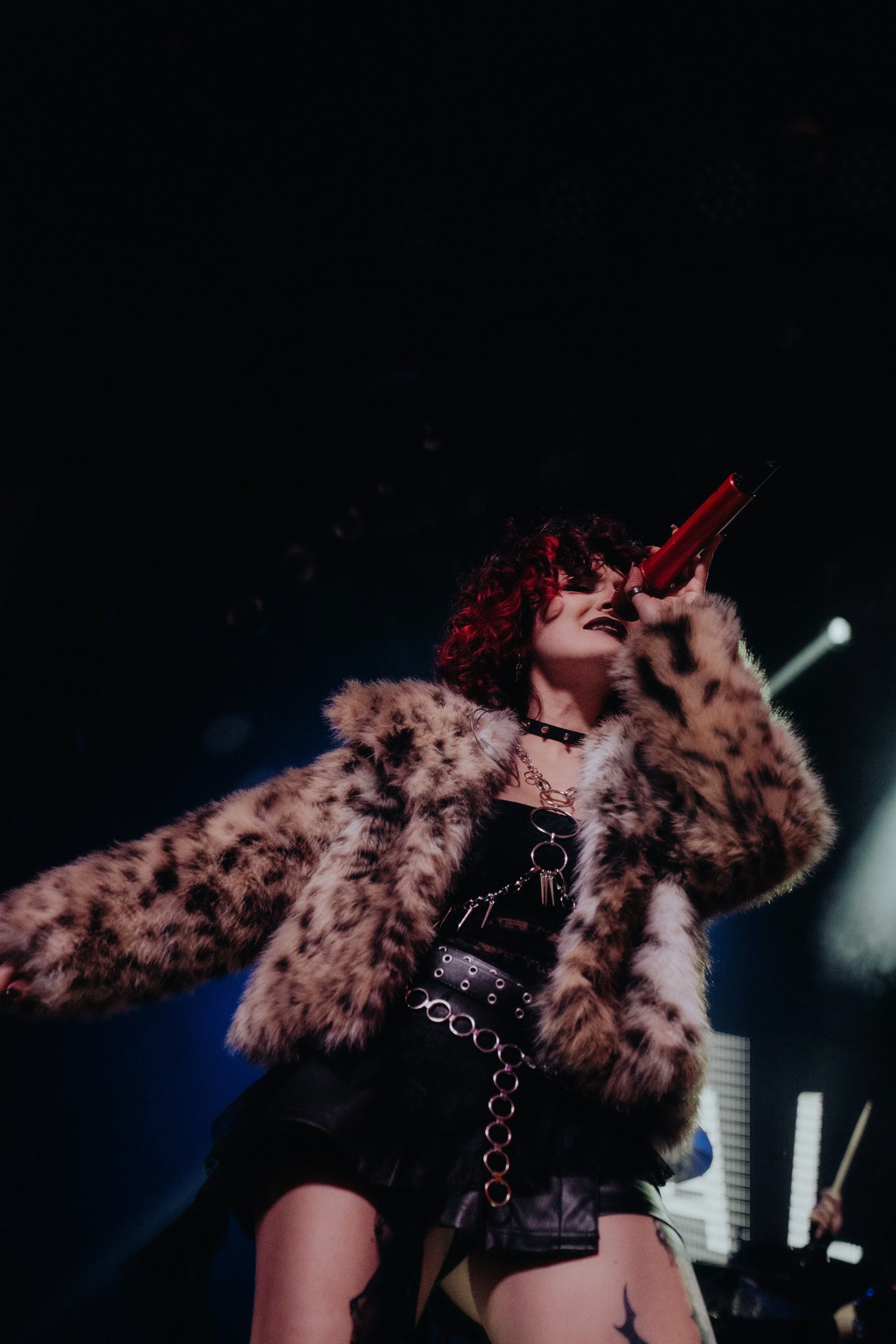 A woman with curly hair, wearing a leopard print fur coat and black choker, singing or speaking into a red microphone on stage with lighting effects.