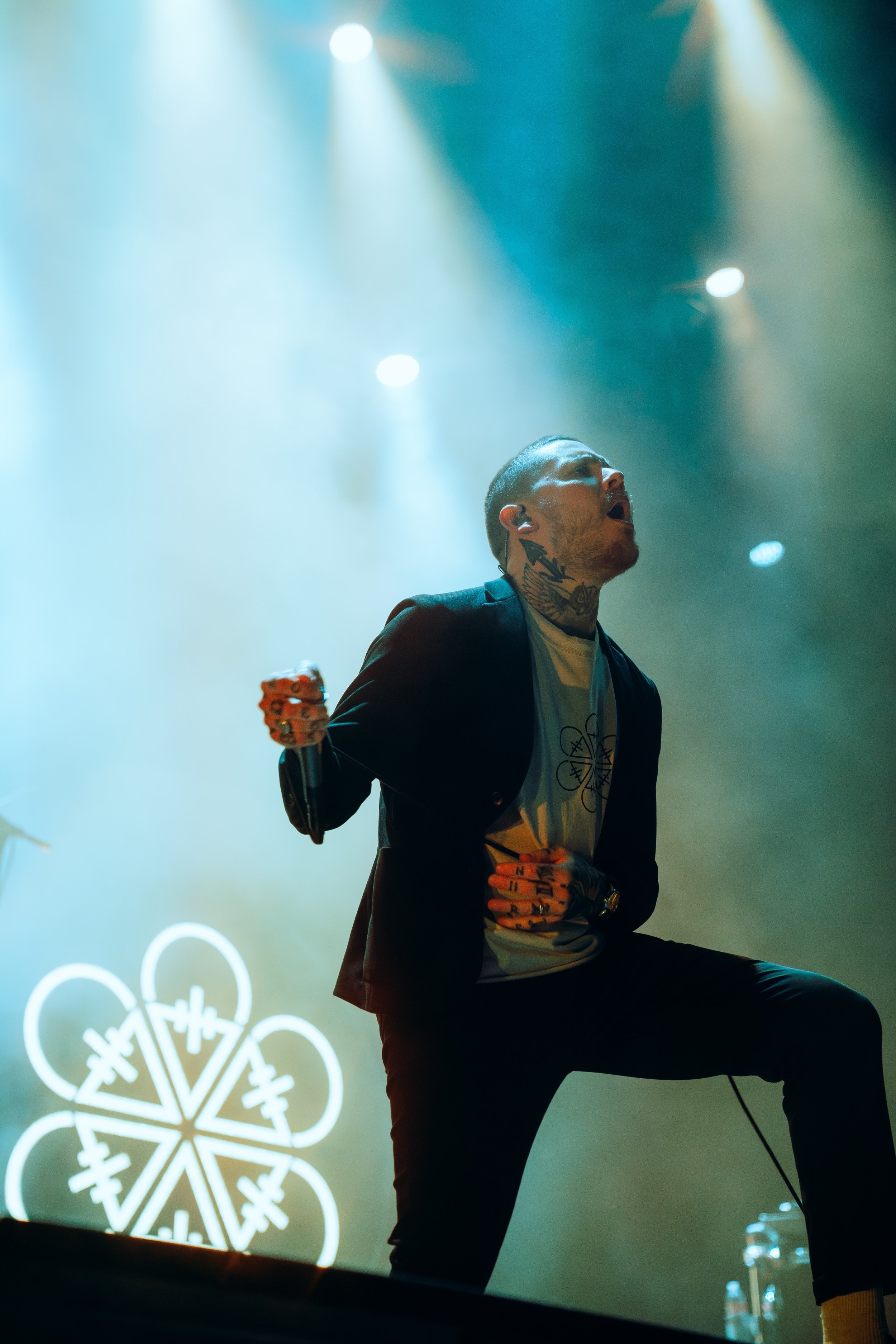 A male singer passionately performs on stage with stage lights shining behind him, wearing a suit jacket over a graphic t-shirt, with tattoos visible on his neck and hands.